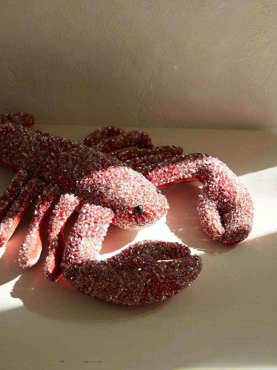 A lifelike lobster sculpture made of red and silver beads sits on a white surface. Sunlight from the left highlights the intricate details of this luxurious artwork.