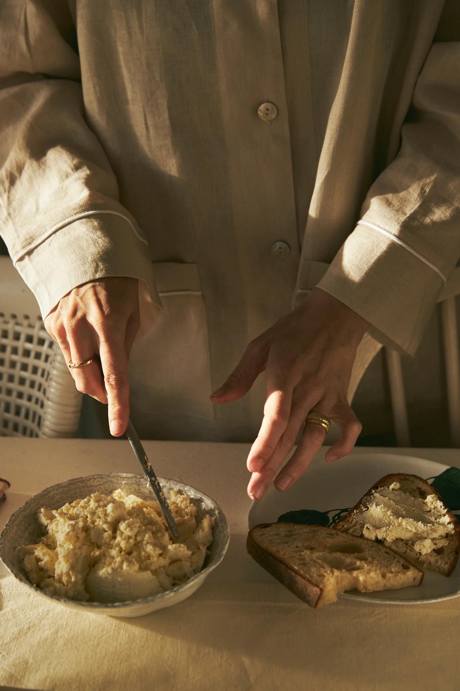 Person in beige spreading cream on bread. Bowl and extra slices nearby. Dimly lit scene on light tablecloth suggests intimate breakfast.