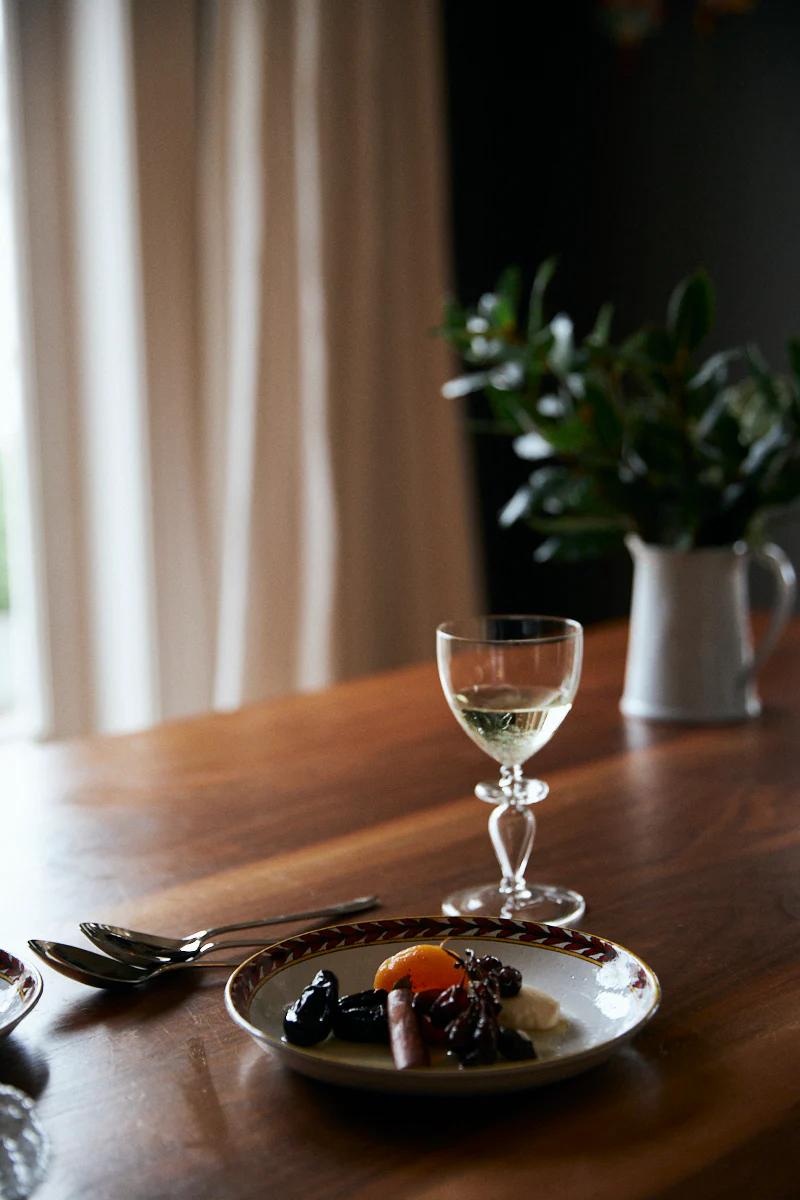 Wooden table with fruit plate, utensils, and white wine glass. White jug with foliage in background.