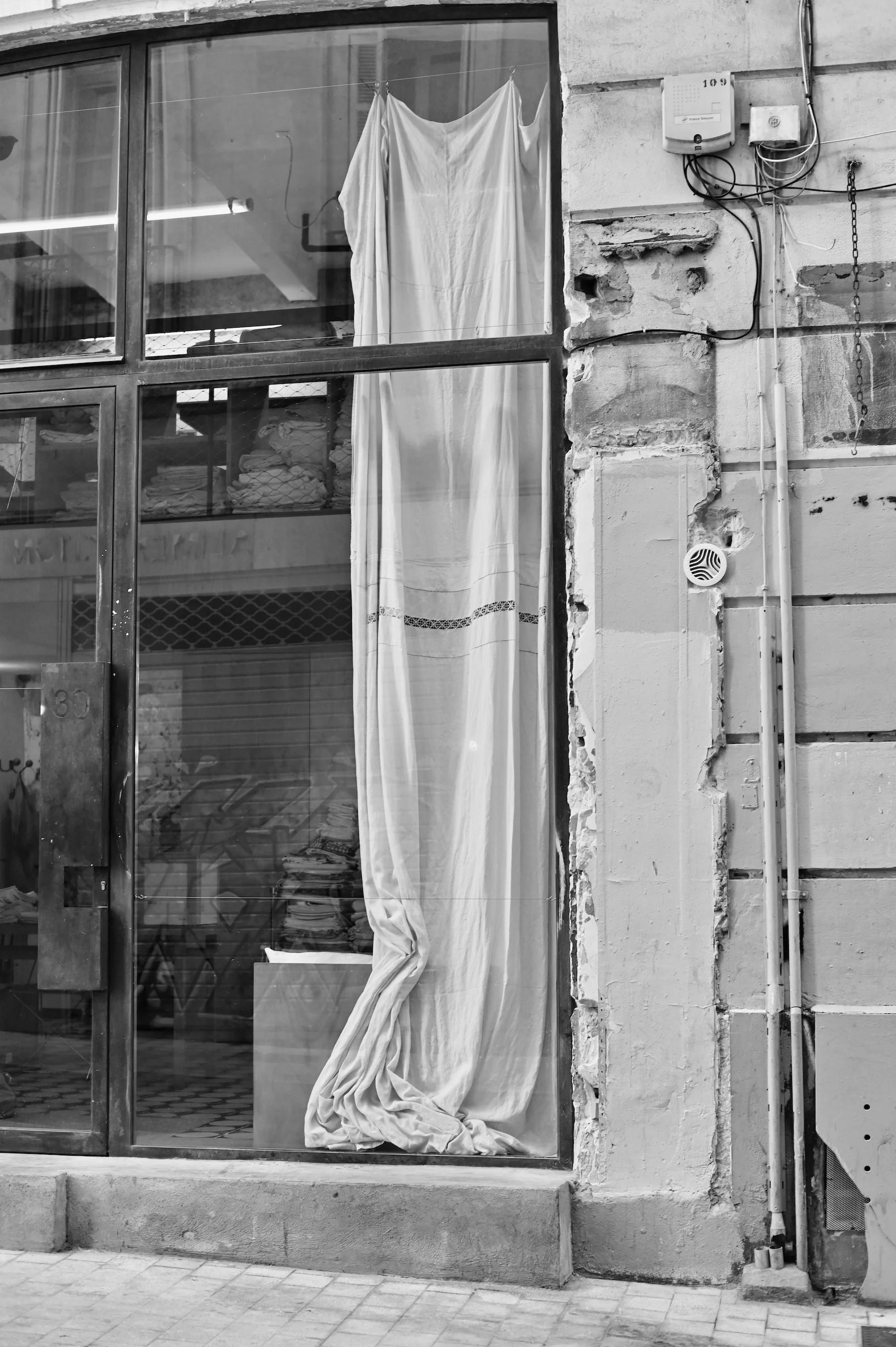 Black and white photo of storefront with large window. White curtain inside, weathered wall with exposed pipes outside.