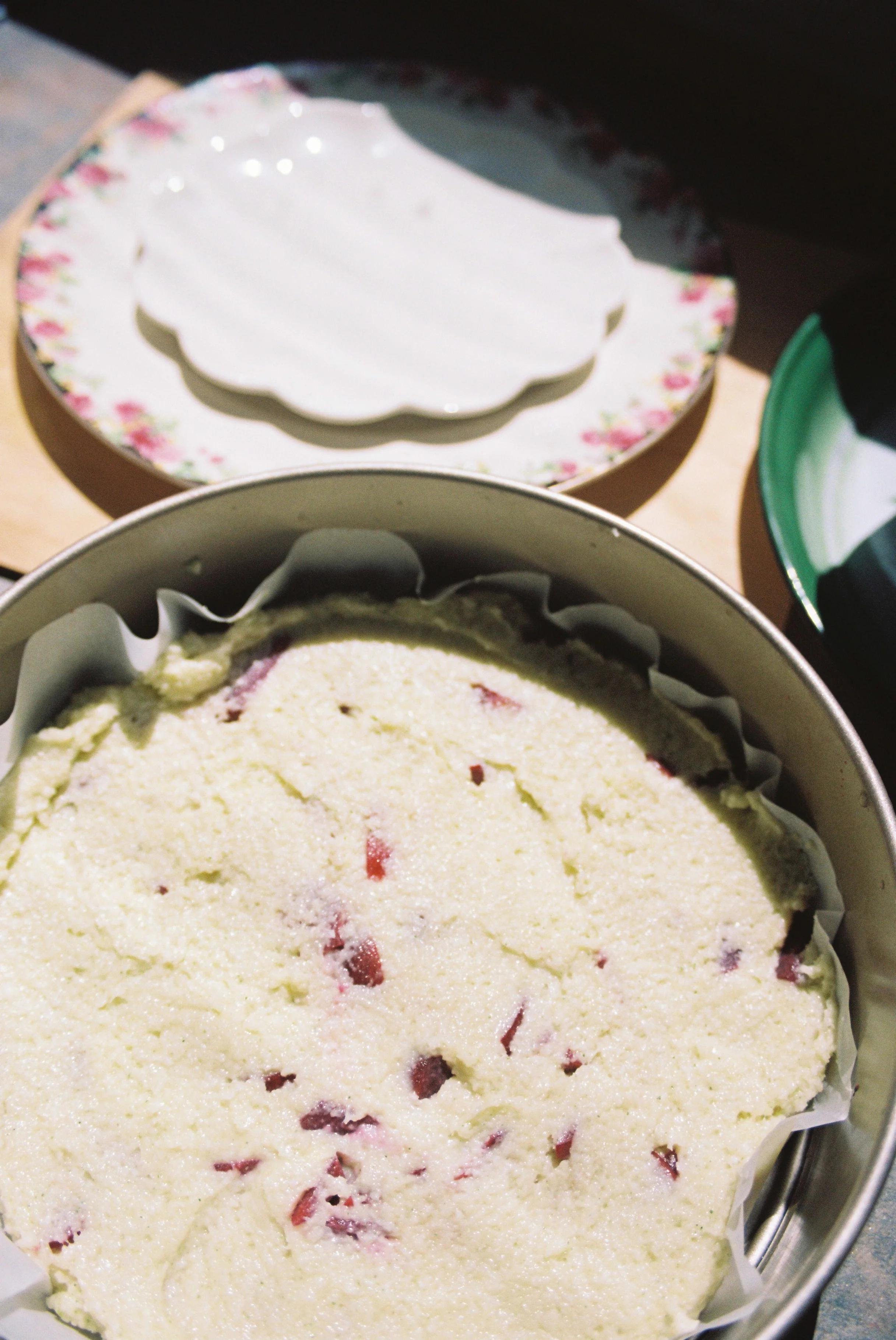 Close-up of unbaked fruit-studded cake batter in lined pan. Floral plate in background. Bright, sunlit scene.