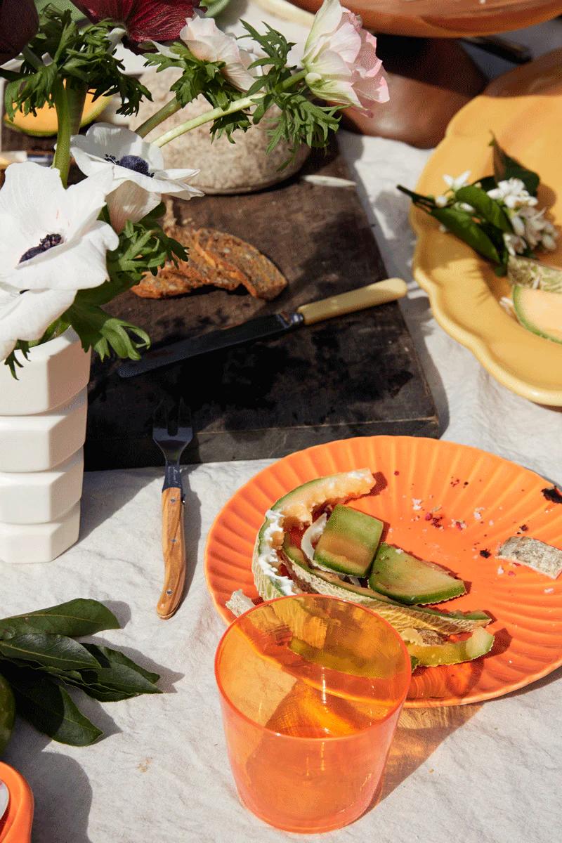 - A picnic table with sliced bread, a half-eaten sandwich, an orange cup, and a white vase with red flowers, surrounded by greenery.