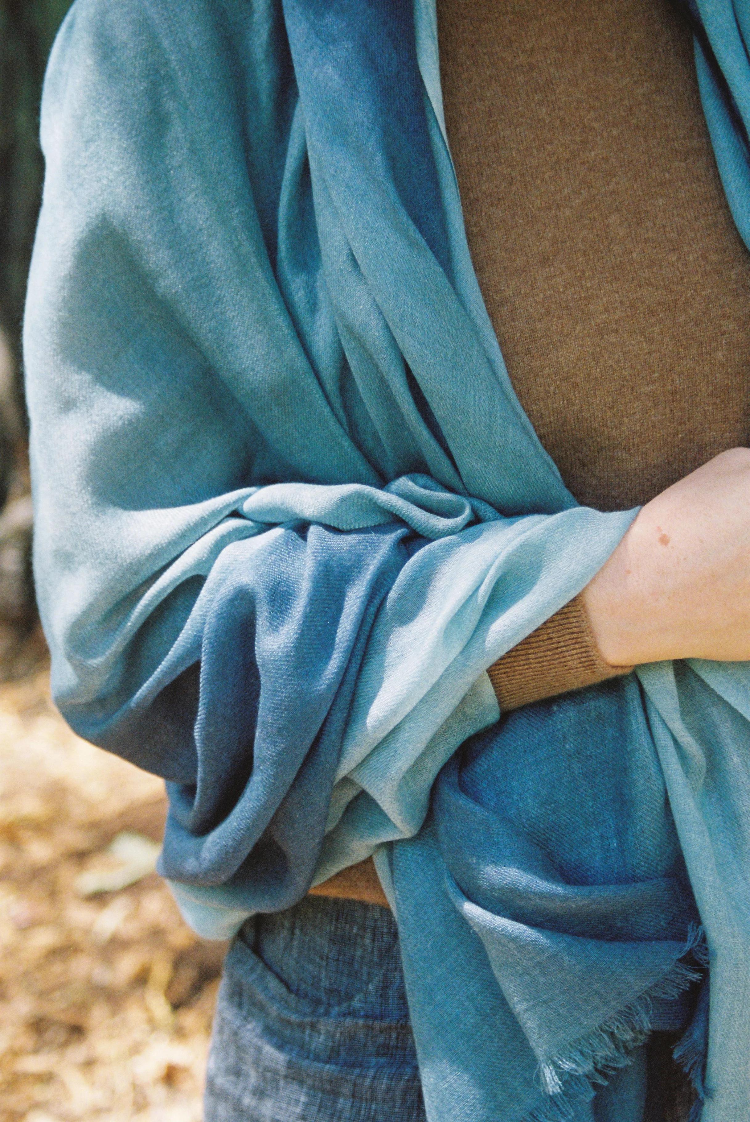 Person in brown top wrapped in gradient blue scarf. Arms and torso visible, holding scarf. Outdoor setting.