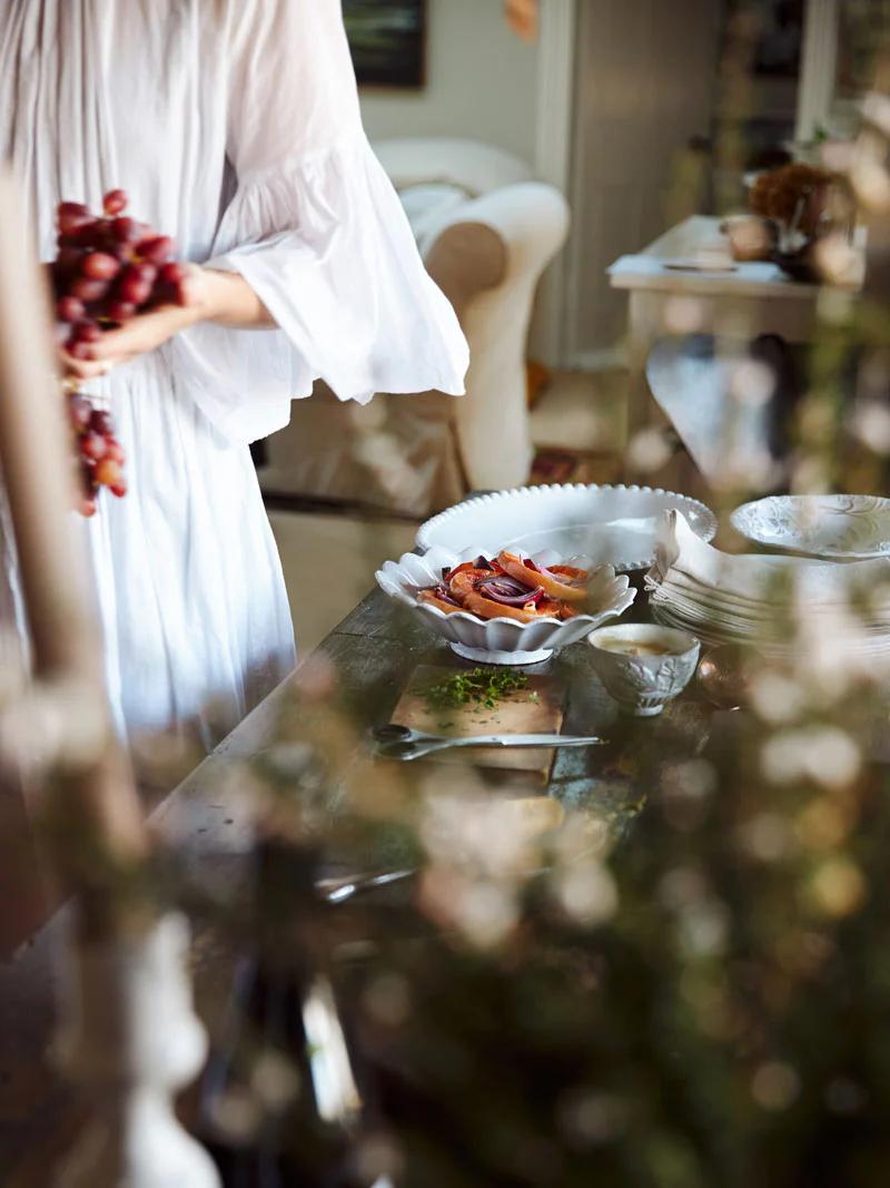 Person in white dress holding grapes near table with fruit bowl. Cozy, softly lit room setting.