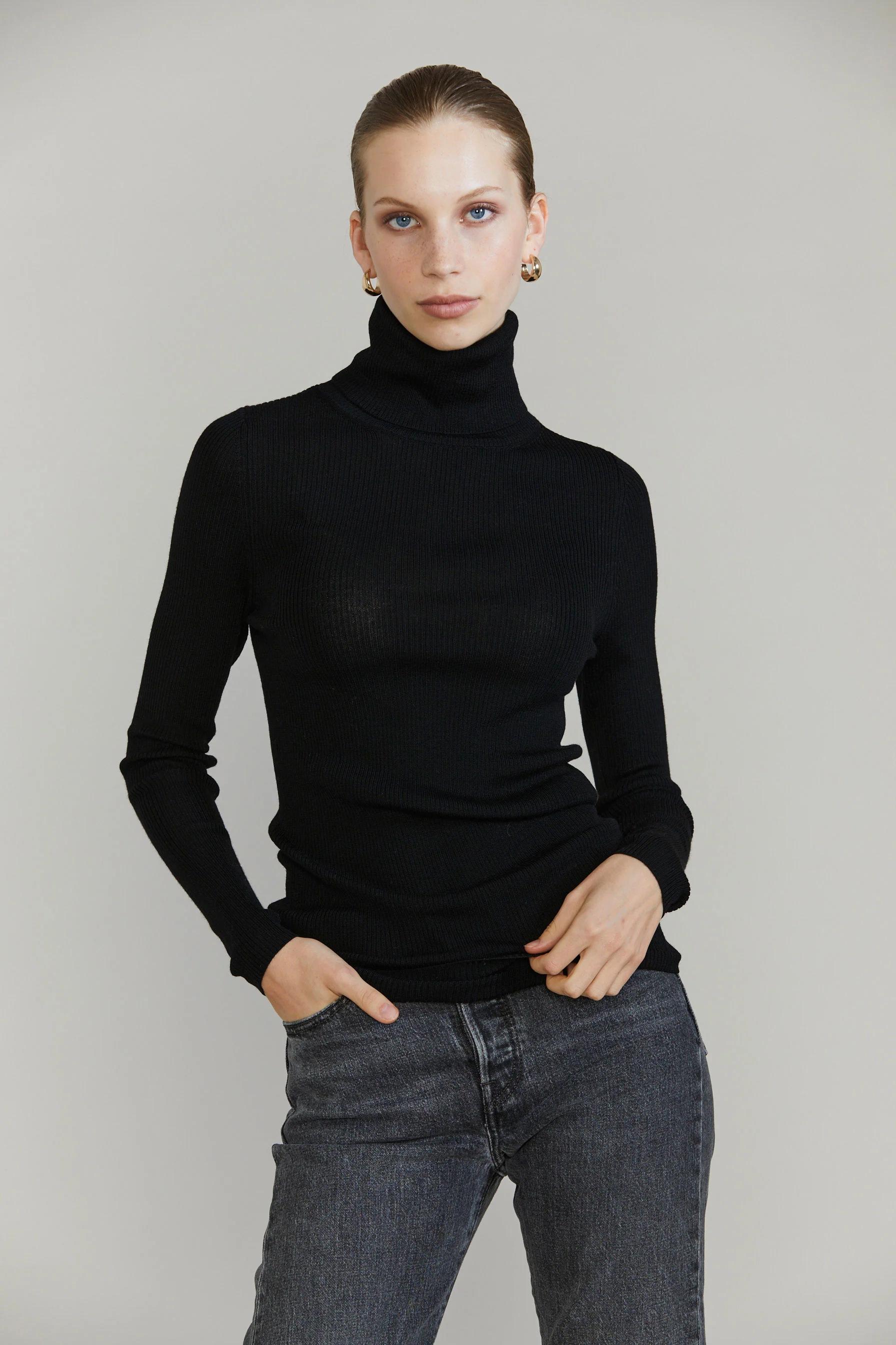 Person in black turtleneck sweater and grey jeans. One hand in pocket. Light-coloured background.