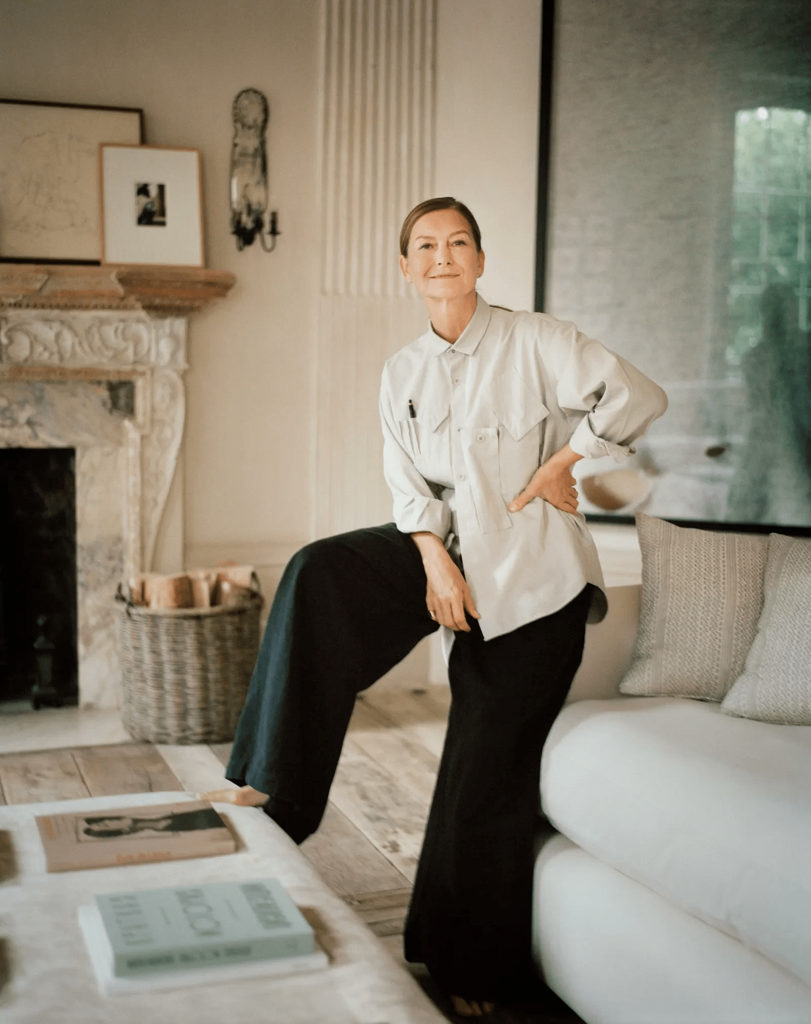 Woman in living room, one foot on couch. Neutral palette, marble fireplace, artwork, coffee table.