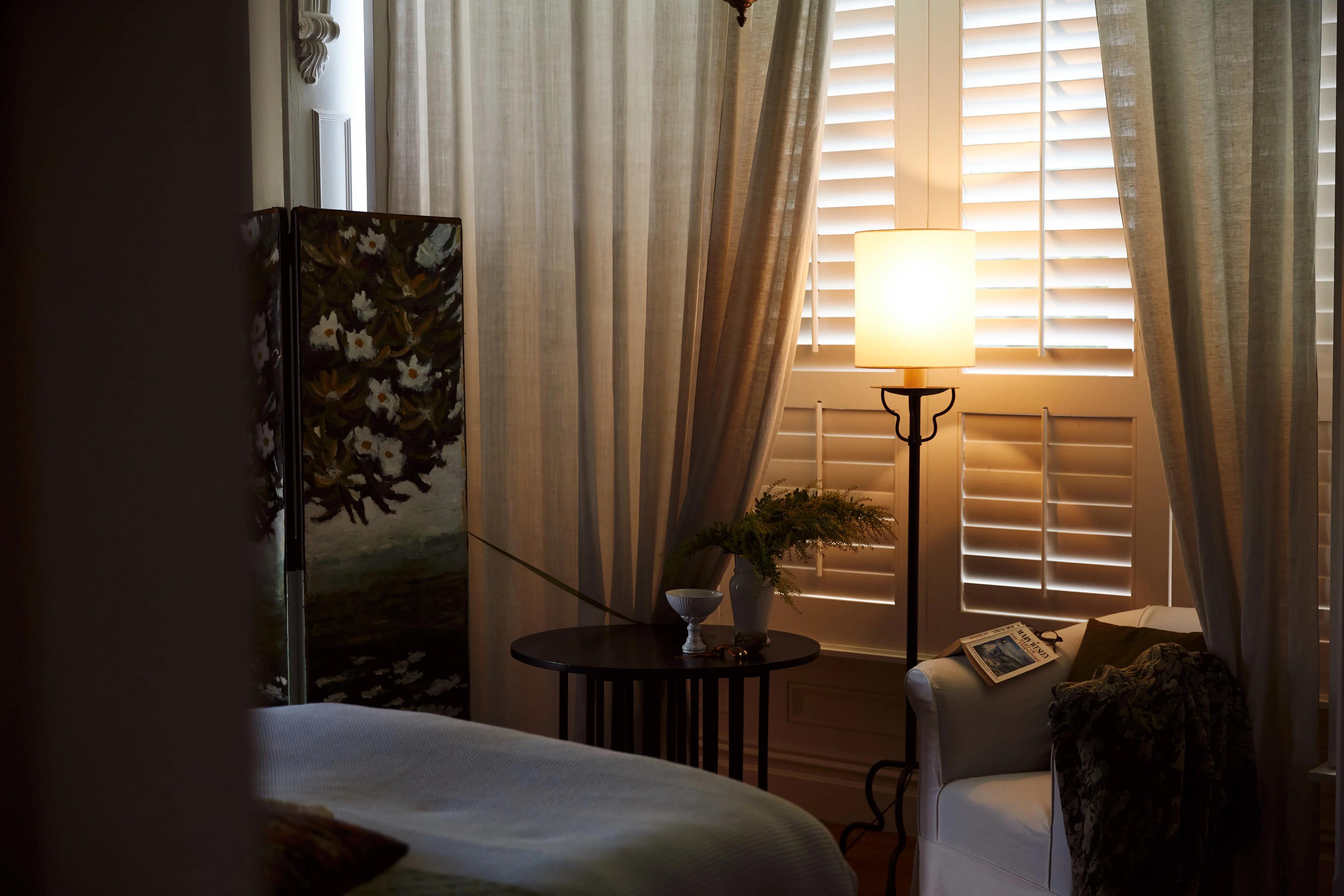 A living room with white shutters and warm lighting from a floor lamp. A small table holds a plant and dish, adding to the room’s inviting, nature-inspired design.