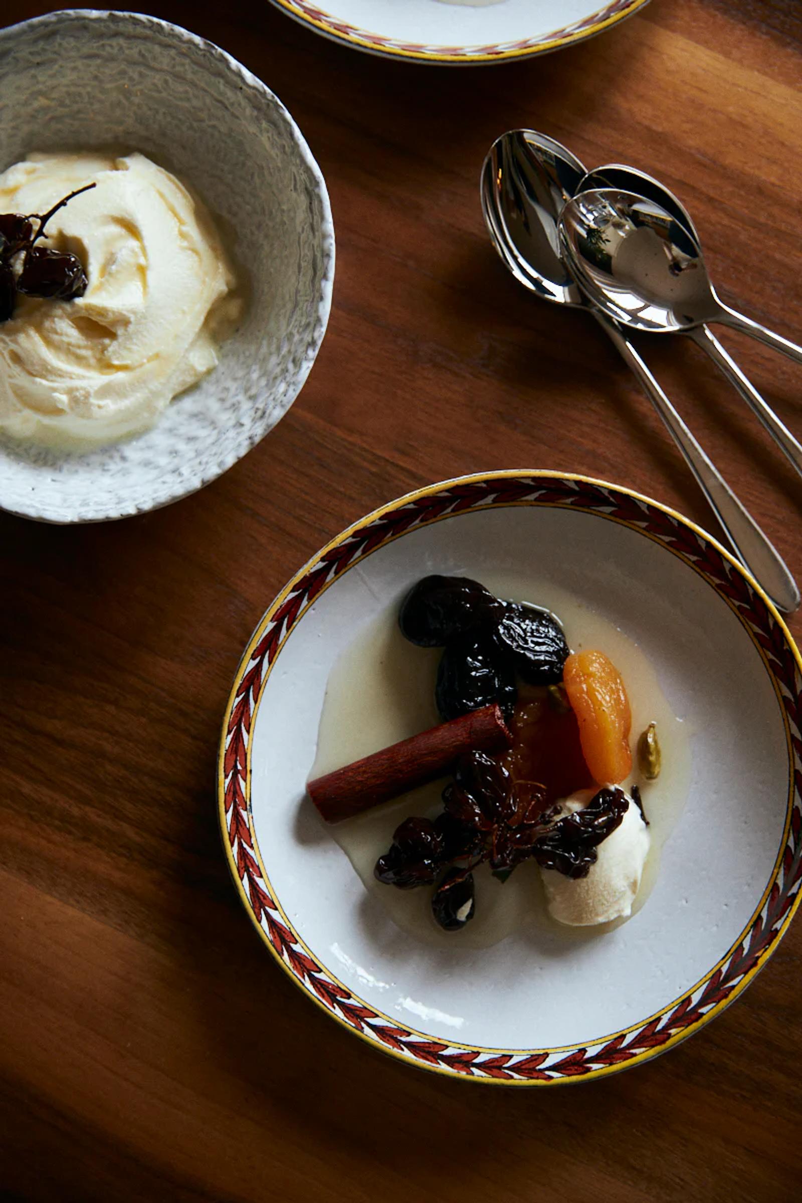 Bowl of ice cream beside plate of dried fruits in syrup with cream. Two spoons on wooden surface.