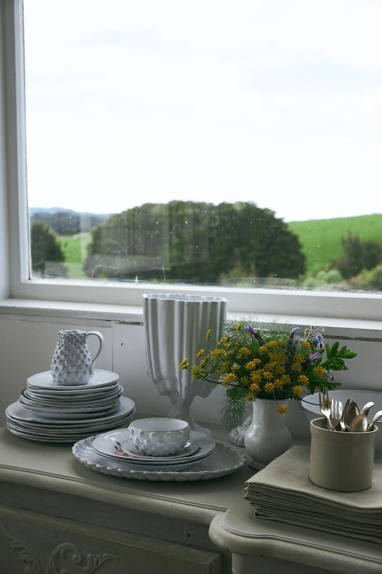 Gray plates and pitcher with tall vases and flower bouquet. Cutlery wrapped in napkins nearby. Window shows outdoor view.