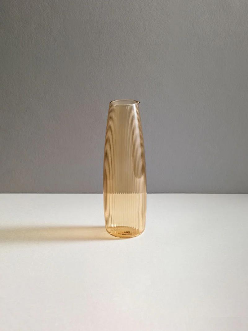 Tall, amber glass vase with ribbed texture on light grey surface. Dark grey background emphasises R+D.LAB's sleek, minimalist design approach.