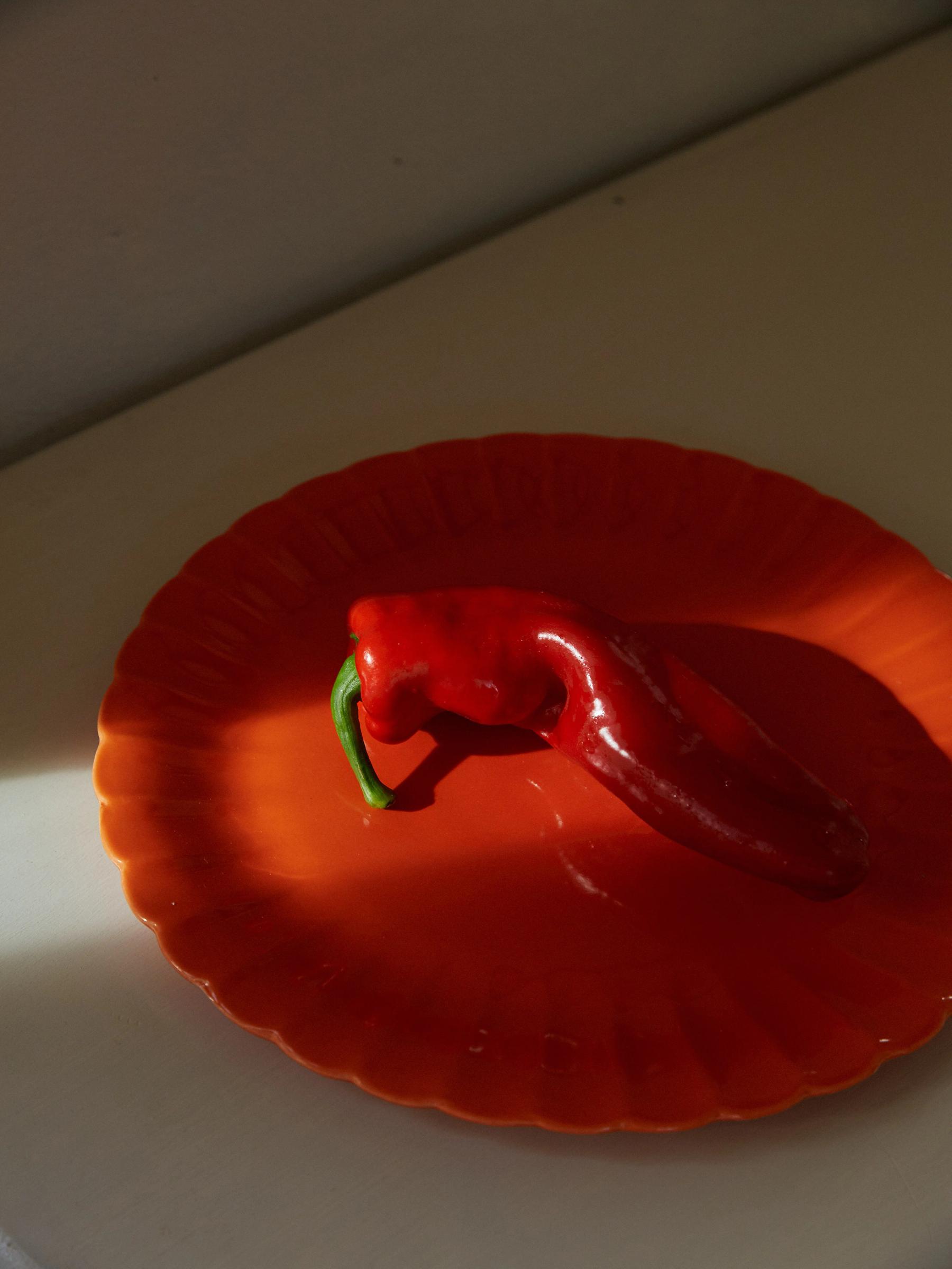 Red chilli pepper with green stem on orange Rachel Carley plate. Natural light creates soft shadows; background blurred.