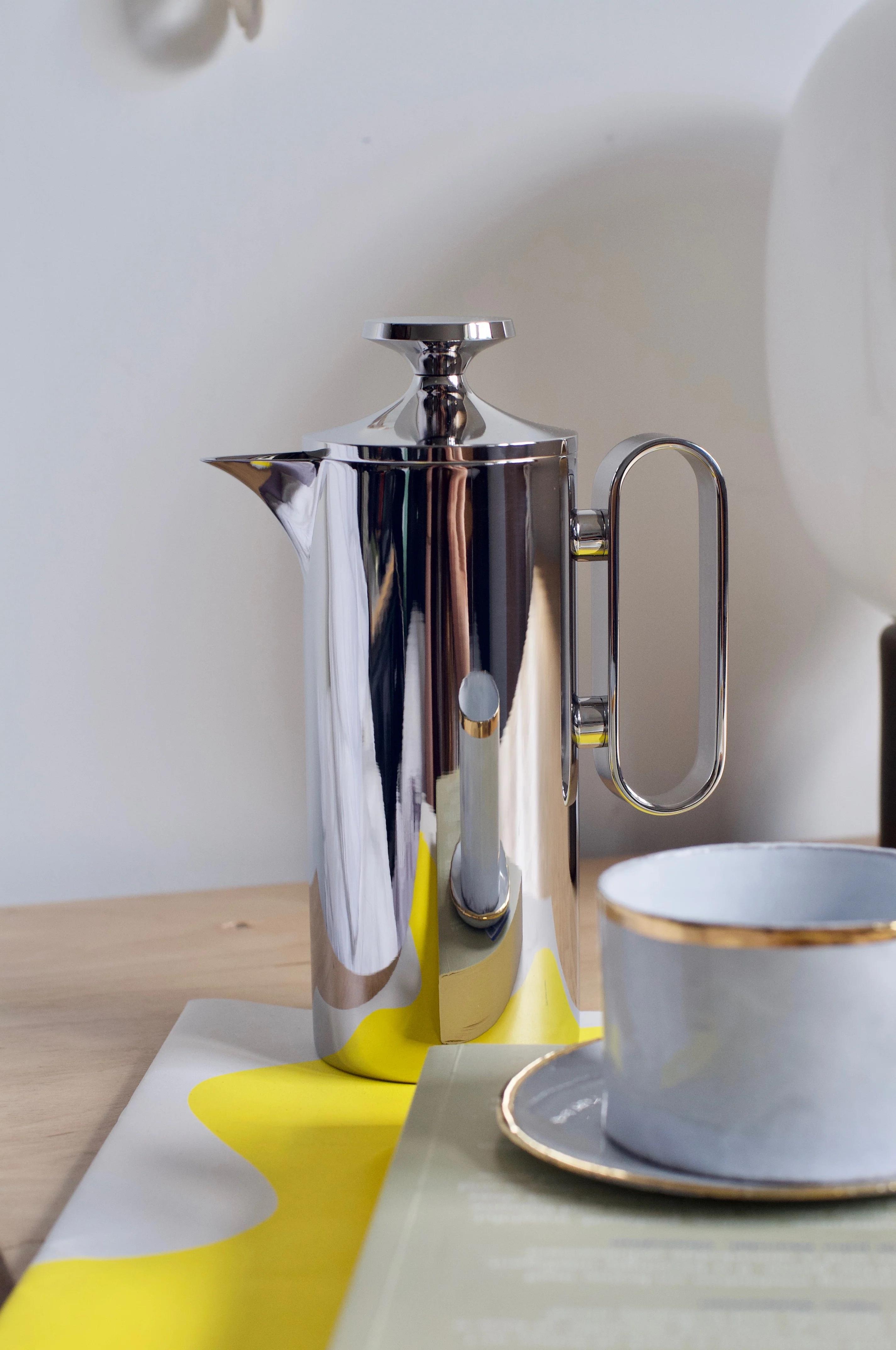 Shiny David Mellor cafetière on wooden table beside white, gold-rimmed cup and saucer. Yellow-white patterned cloth and book corner visible.