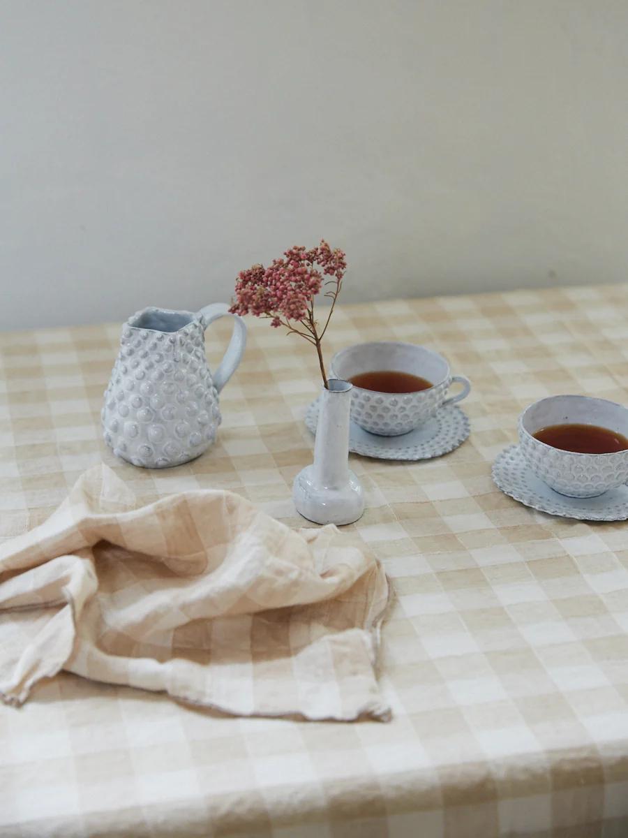 Checkered tablecloth with ceramic pitcher, flower vase, teacups on saucers, striped beige cloth.