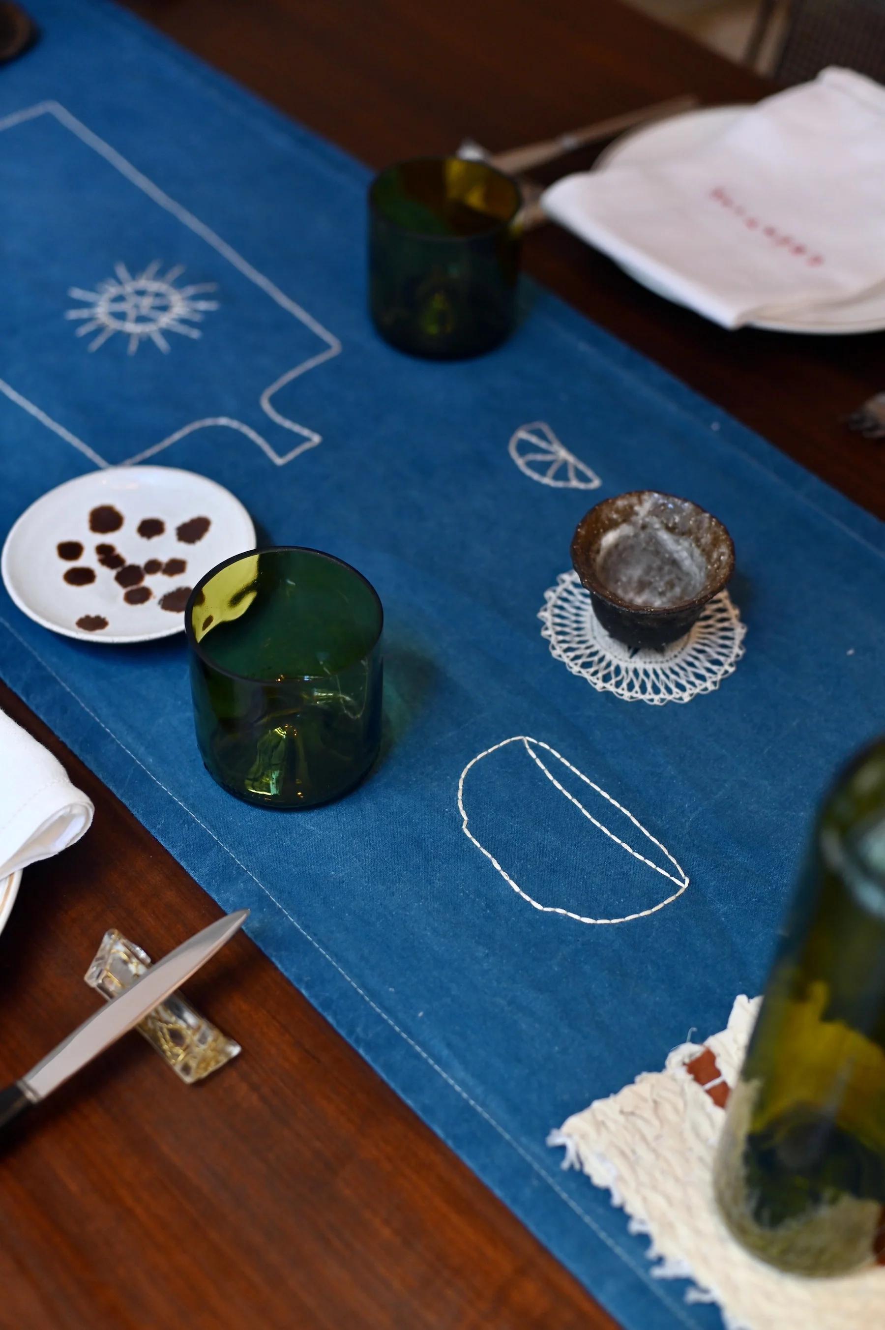 Table set with blue runner, green glasses, white dish, textured bowl, knife, and folded napkins.