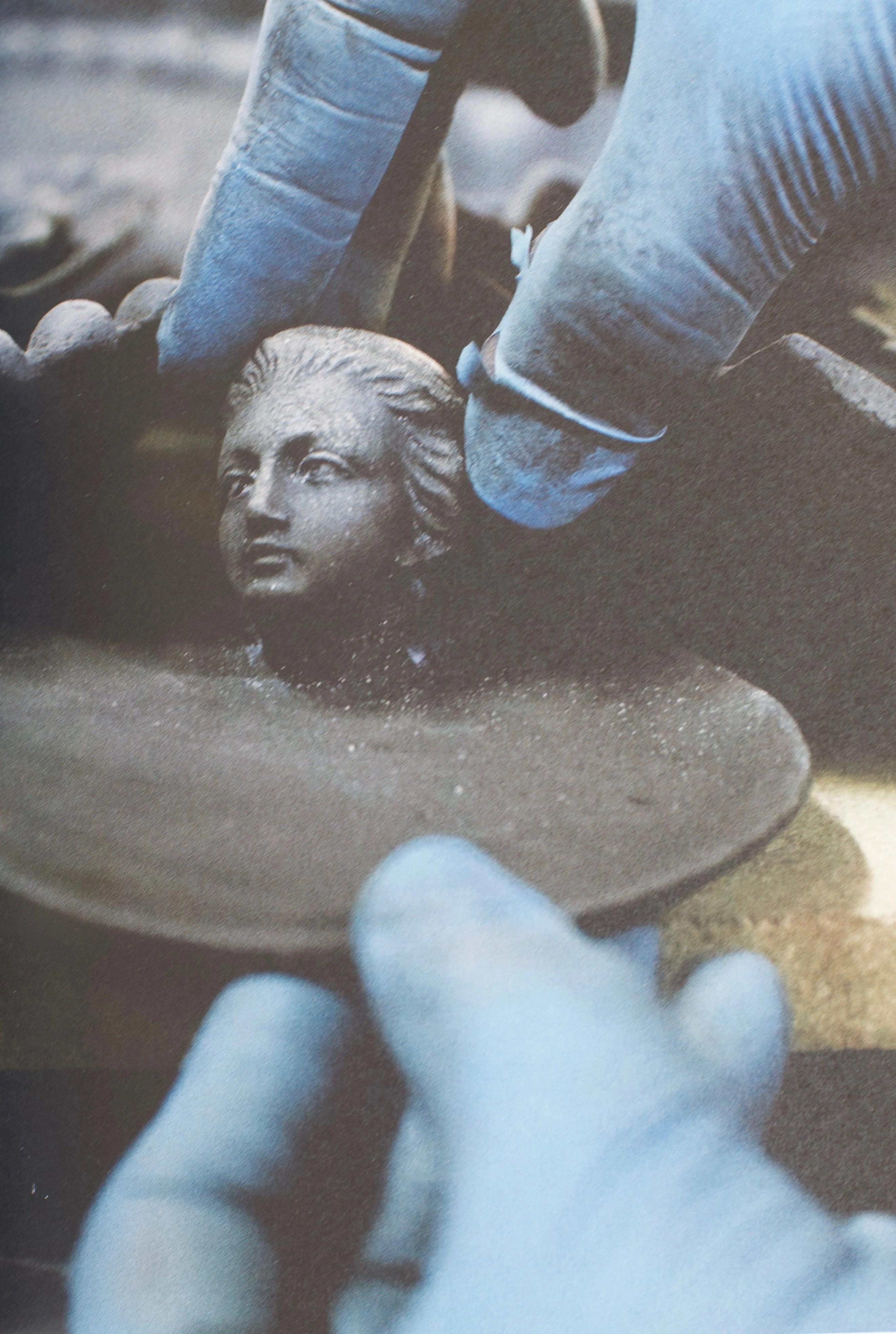 Gloved hands work on a small, detailed clay sculpture of a human face. The image focuses on the intricate artisanal process.