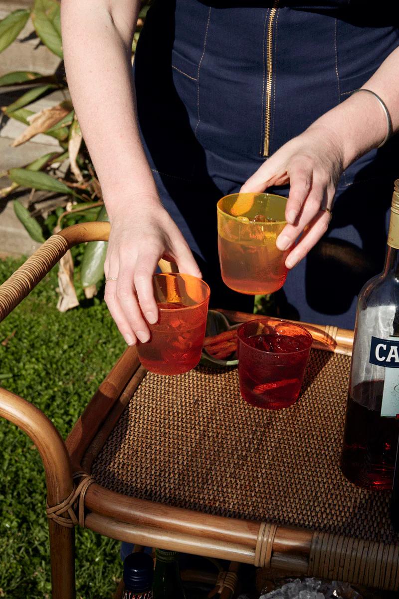 Person arranging colourful drinks on outdoor wicker cart. Campari bottle visible. Lush green foliage surrounds.