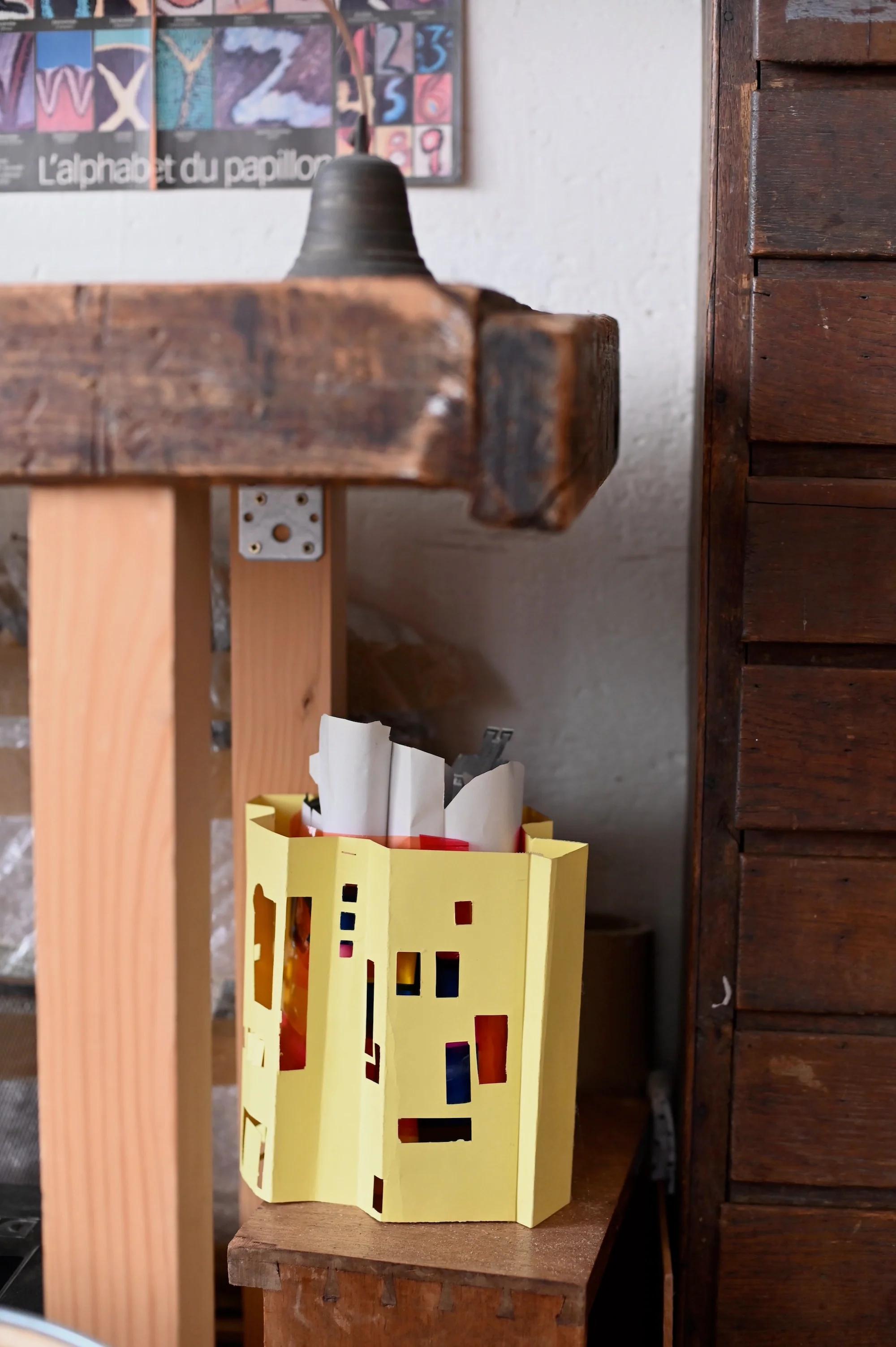 Handmade yellow paper basket on wooden shelf. Filled with paper, next to wooden structure with metal clamp. Colourful chart background.