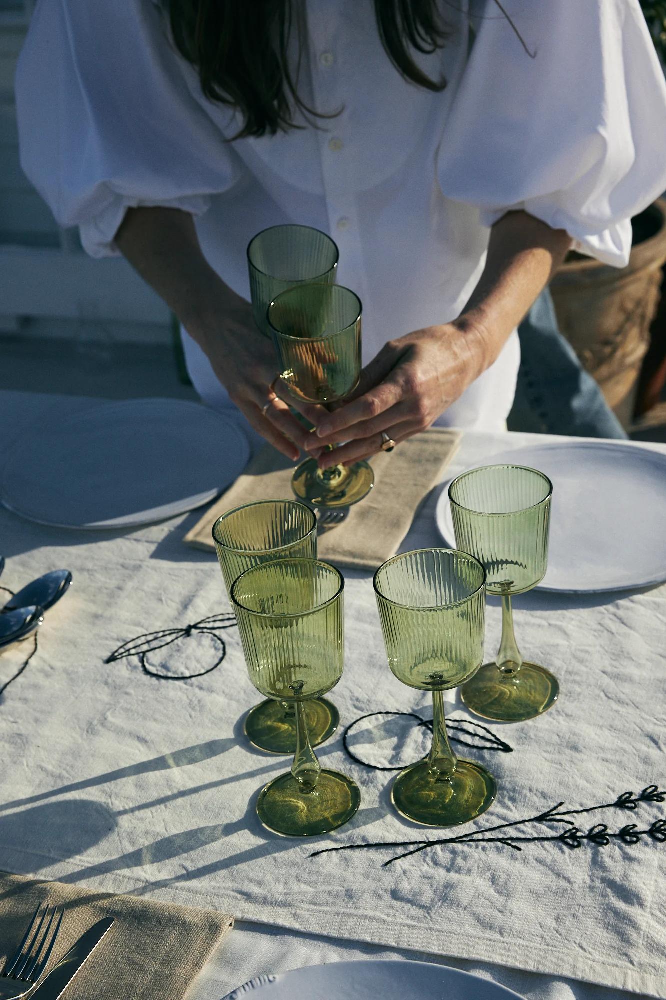 Person arranges green glass goblets on linen tablecloth. Table set with white plates, cutlery, and napkins.