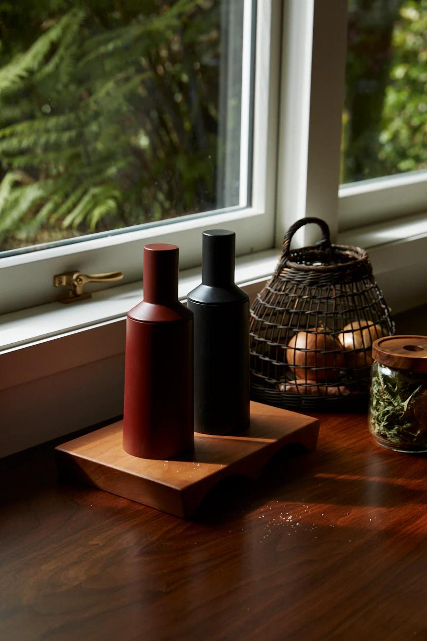 Modern red and black containers on a wooden tray by a window. A rustic basket with eggs and herbs sits nearby, blending contemporary and classic aesthetics.