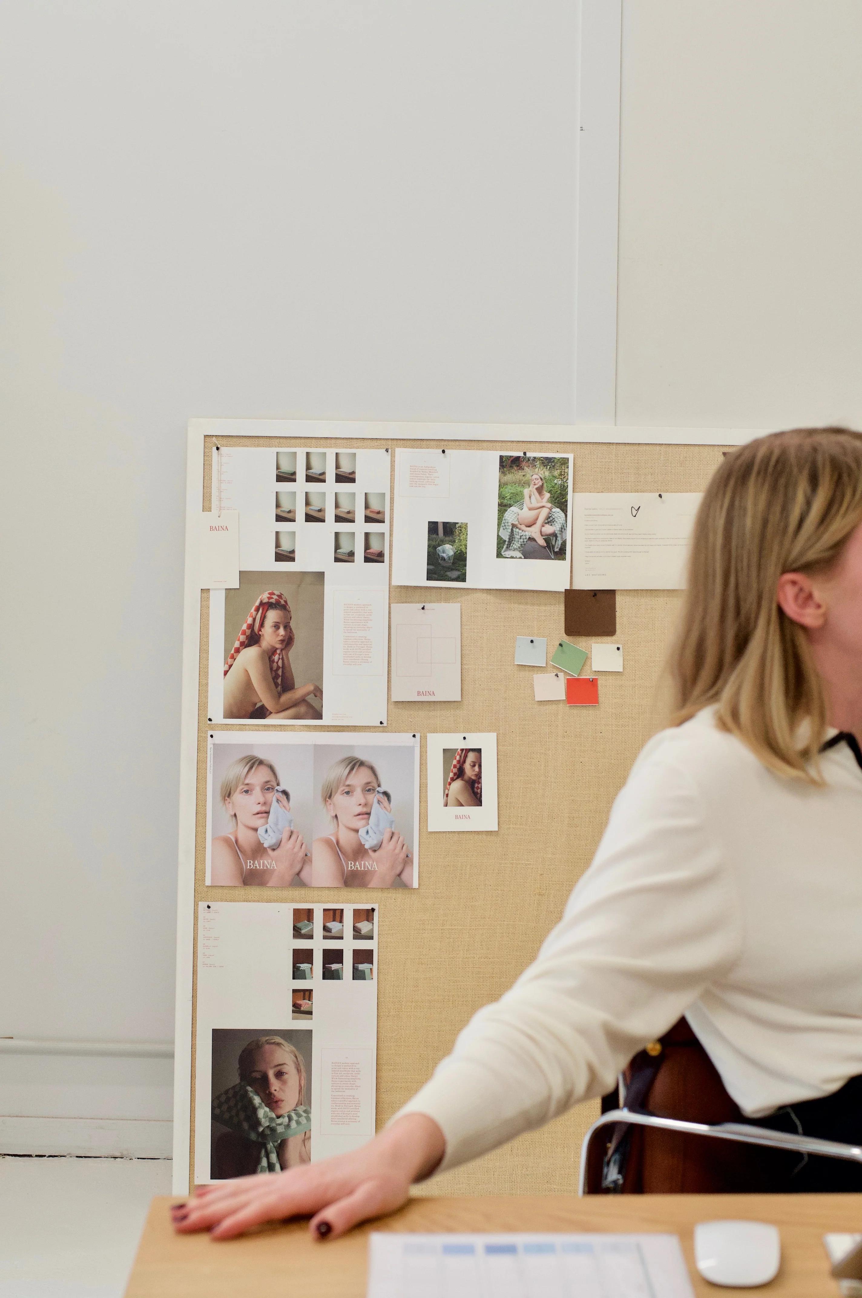 Workspace with mood board. Blonde person in white top at desk. Minimalist background with white walls.