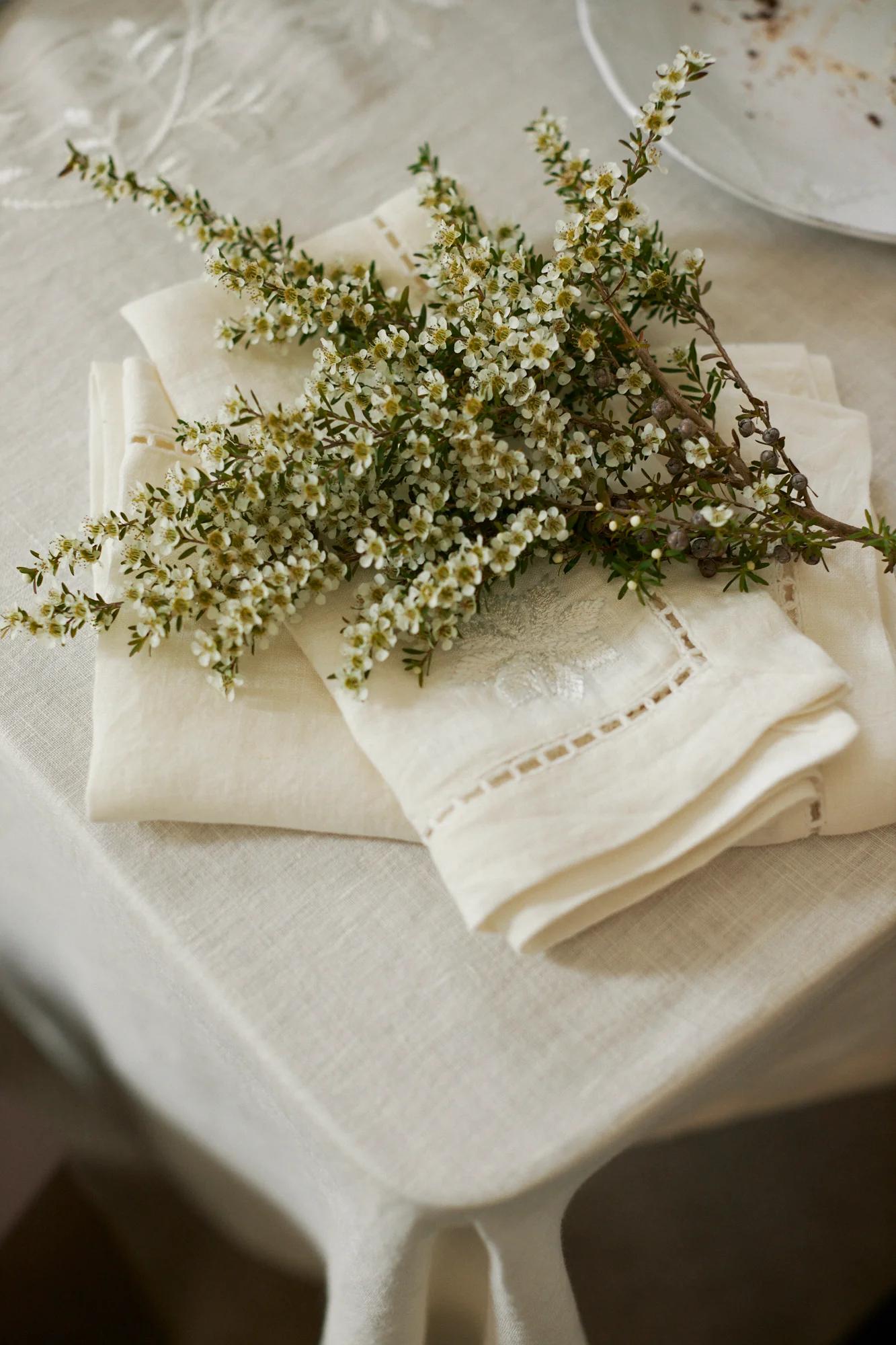 A small bouquet of white flowers rests on folded cream napkins atop a white tablecloth. Part of a white plate is visible, creating an elegant table setting.