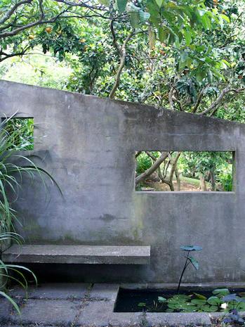 Concrete wall with openings reveals lush garden. Rachel Carley's bench overhangs lily pond. Tall plants enhance the hidden oasis feel.