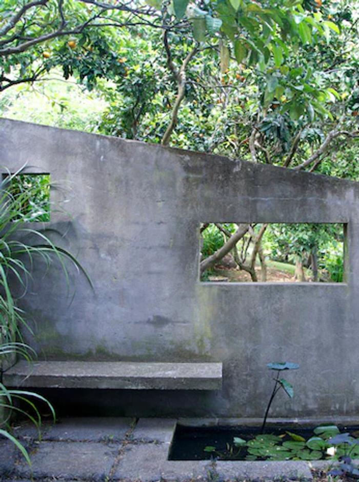 Concrete wall with openings reveals lush garden. Rachel Carley's bench overhangs lily pond. Tall plants enhance the hidden oasis feel.