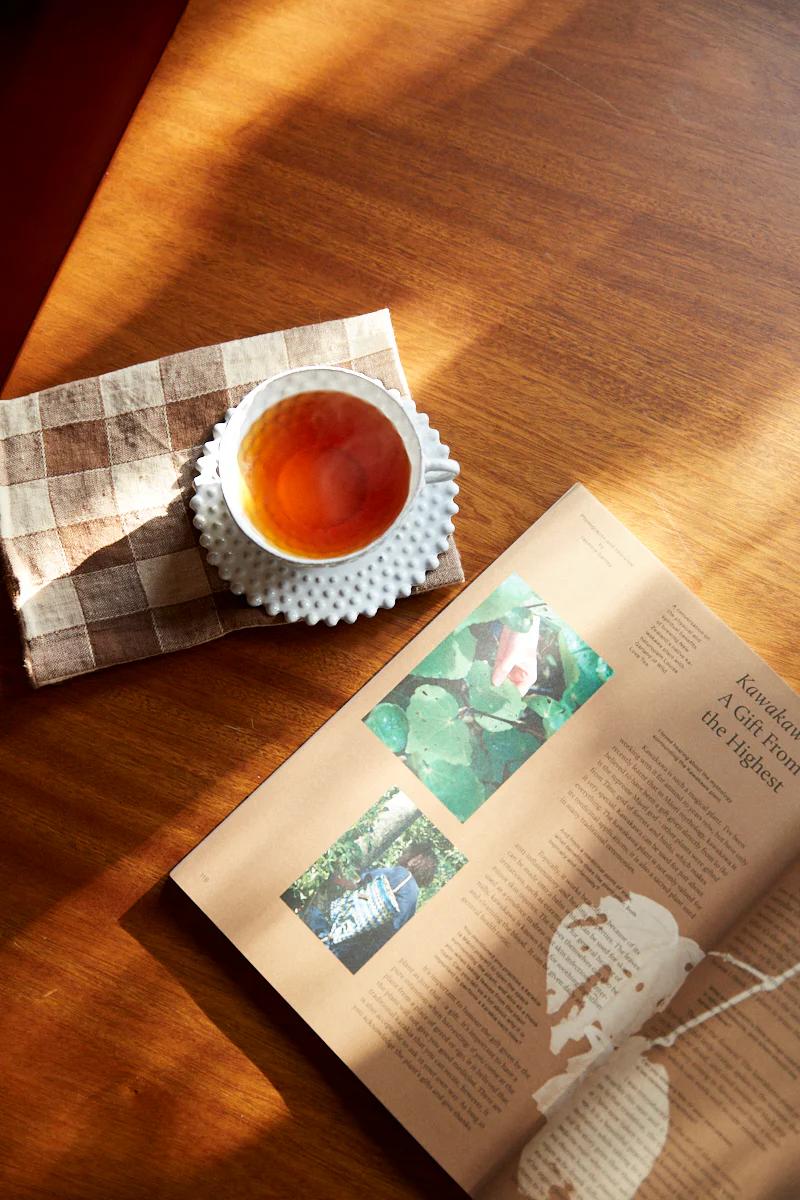 Tea cup on saucer and checkered napkin. Open magazine nearby. Sunlit wooden table.