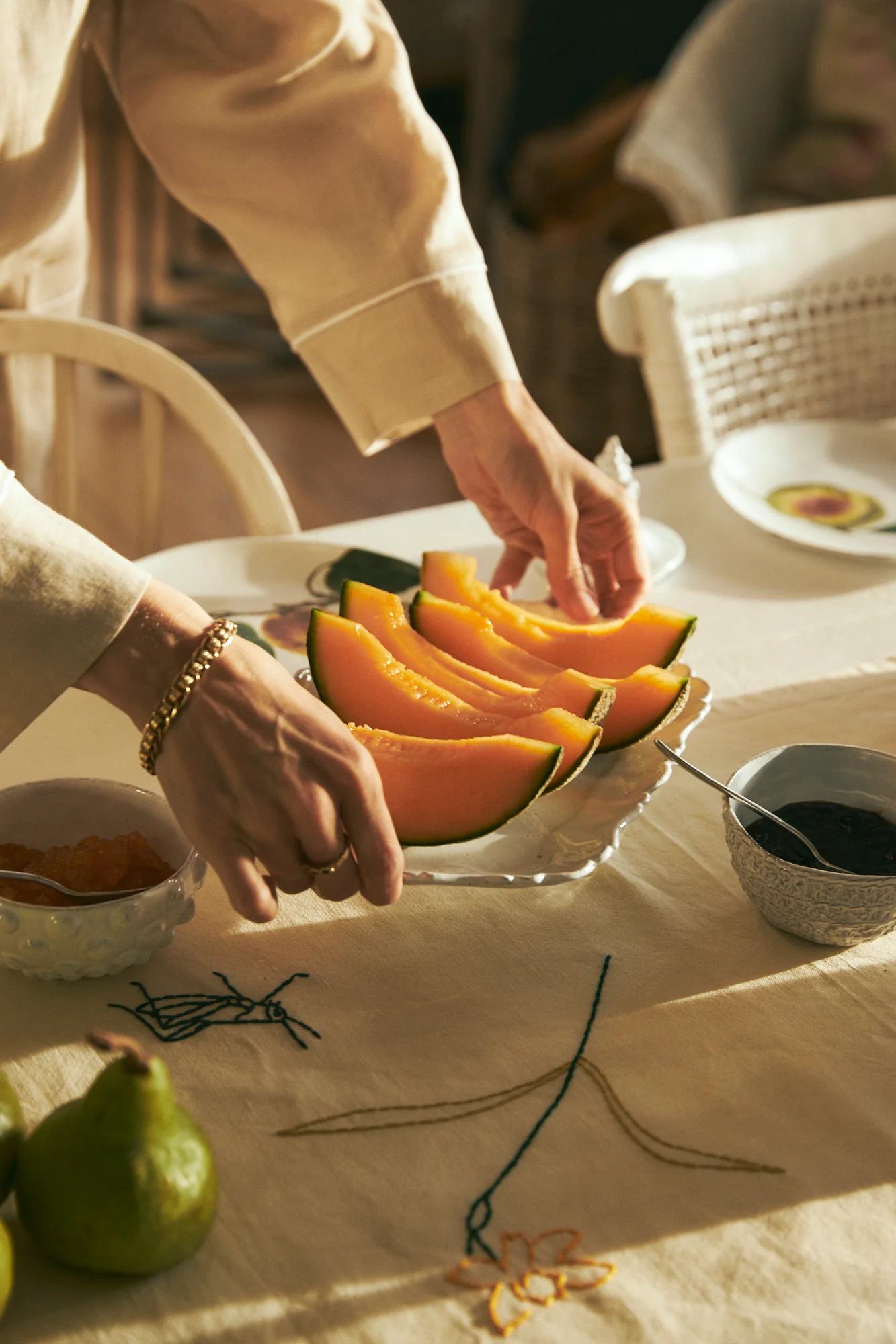 Person arranging cantaloupe plate on embroidered tablecloth. Table set with various bowls and a pear. Captures cosy breakfast scene.