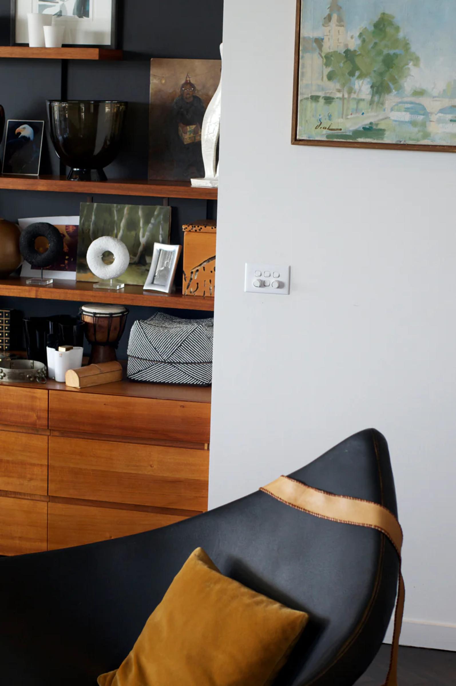 Black leather chair with mustard pillow in foreground. Wooden shelves display art and objects behind. Painting of Sydney hangs above light switch on white wall.