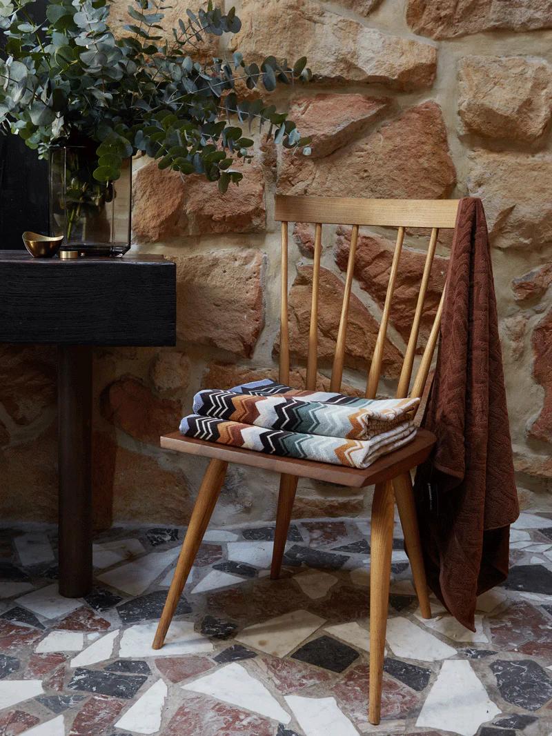 A wooden chair with colourful towels on the seat and a brown towel draped over the back, placed on a patterned tile floor beside a side table and stone wall.