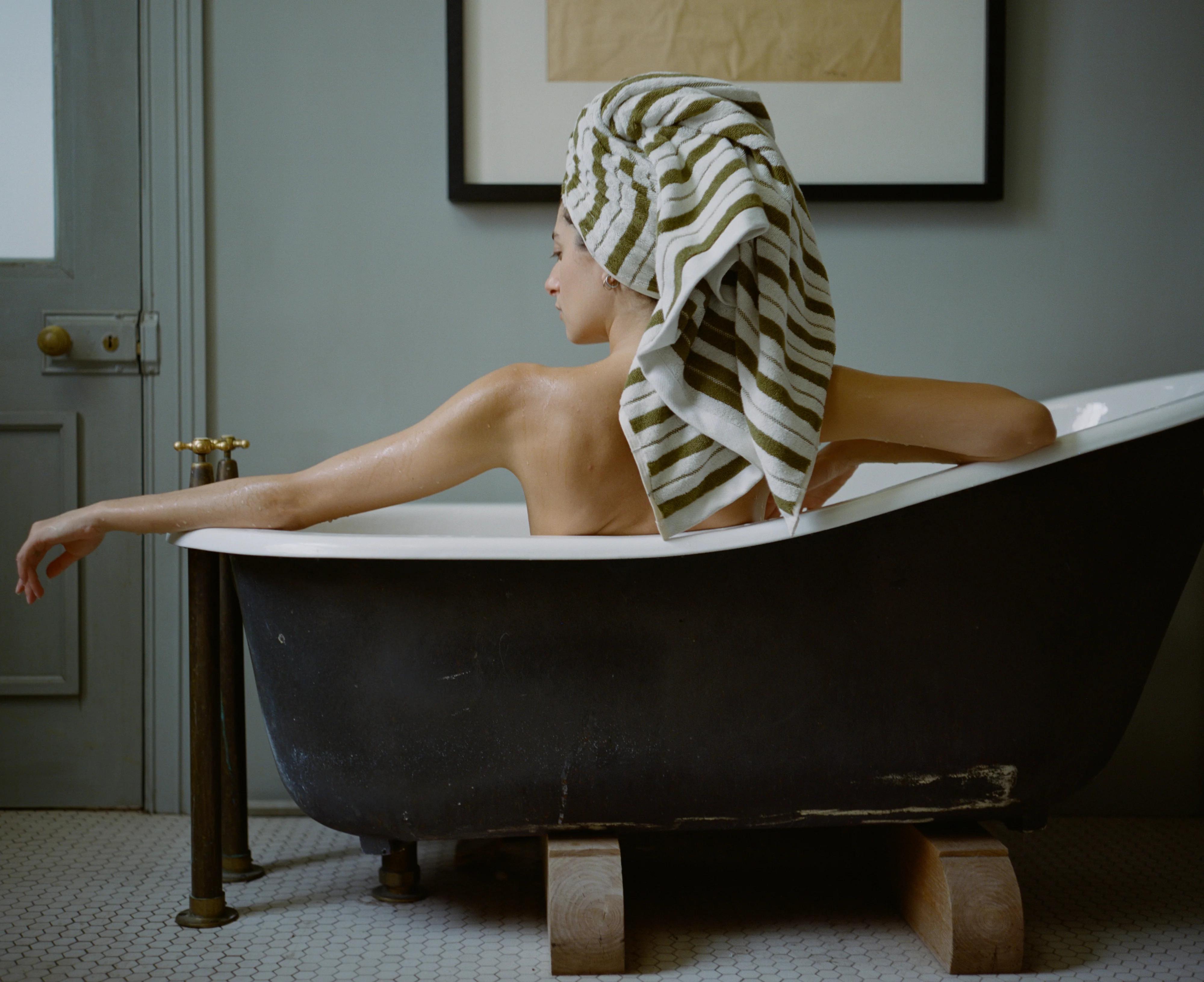 Person with towel-wrapped head in black freestanding bathtub. Tiled floor and wooden door in background.