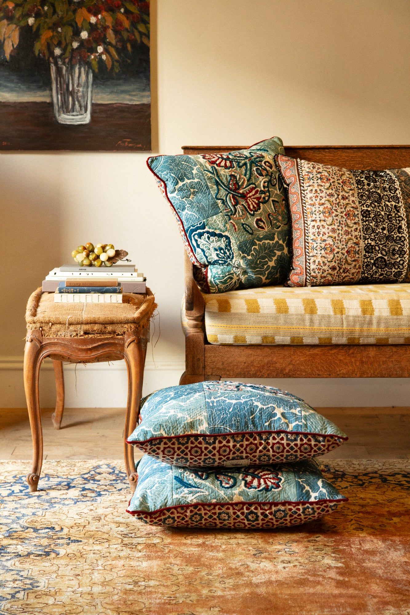 Cosy room with wooden bench, patterned cushions. Side table holds books and grapes. Floor cushions, rug, and floral art evoke British charm.