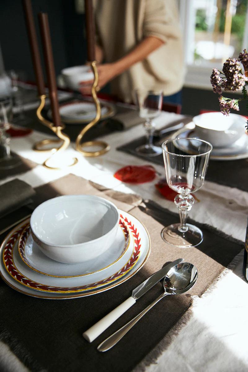 An elegant table setting with white bowl, decorative plate, cutlery, and wine glass. Two dark candles stand tall as someone arranges the table.