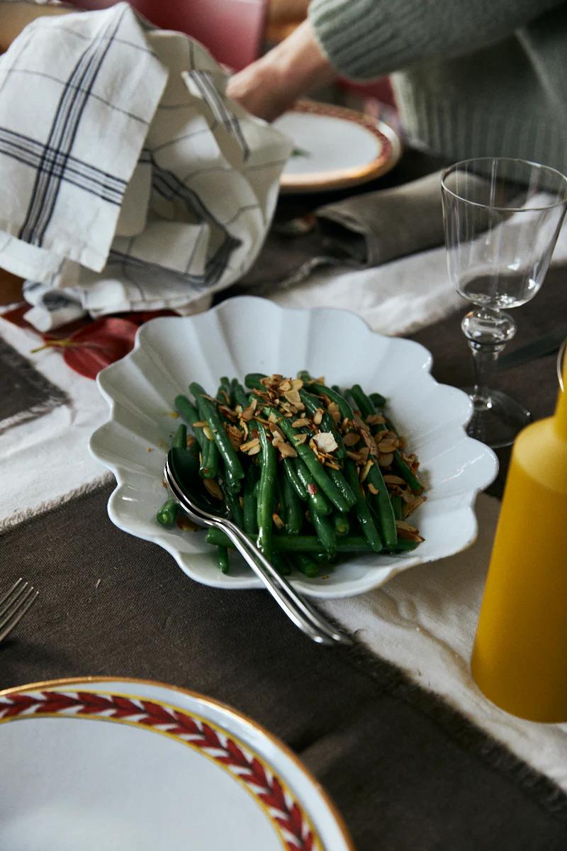 White bowl with green beans and almonds. Plates, glass, cutlery, yellow bottle, and napkin nearby. Person in background.