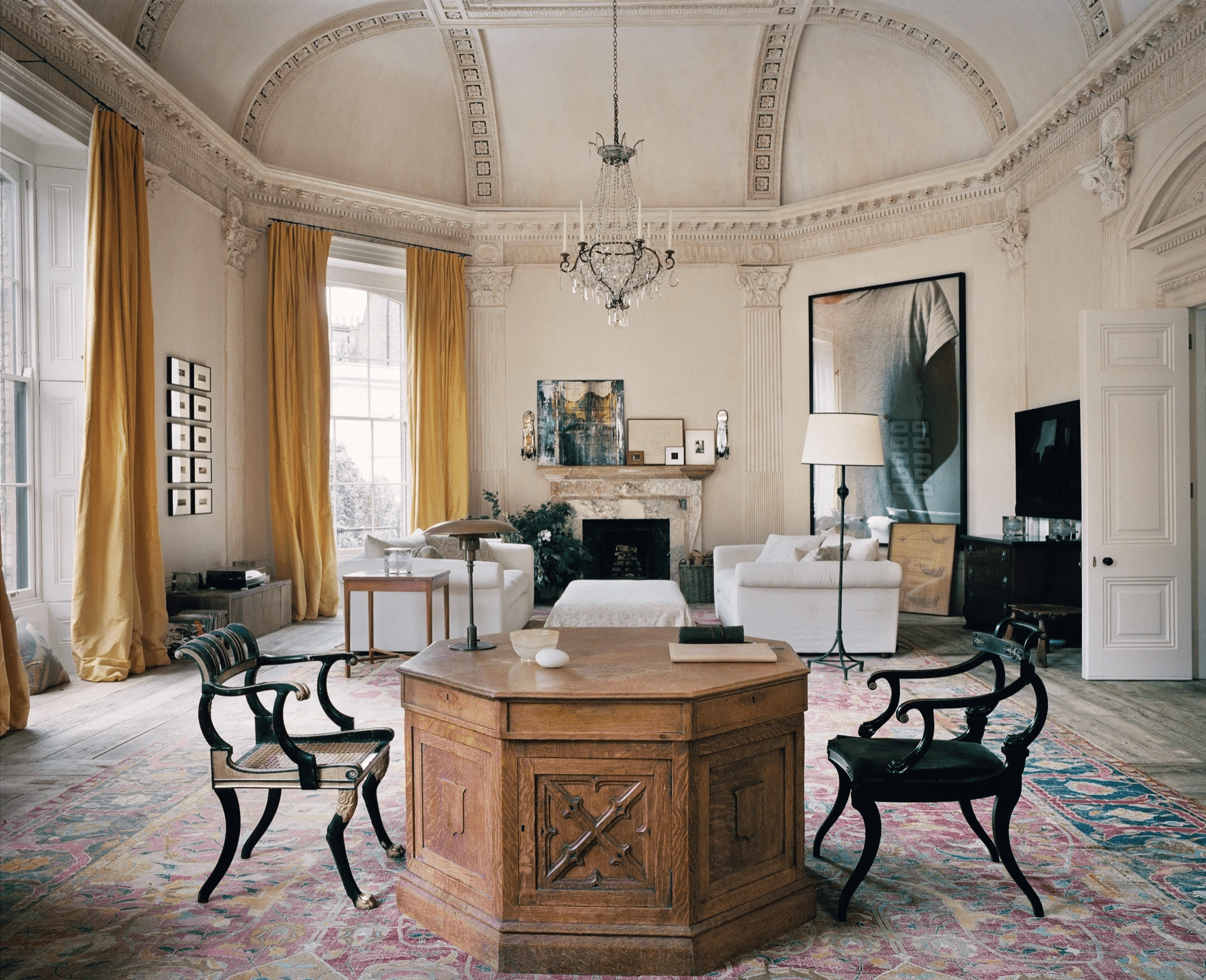 Grand living room with ornate ceiling. Large windows, chandelier, fireplace with art. Mix of classic and modern furniture.