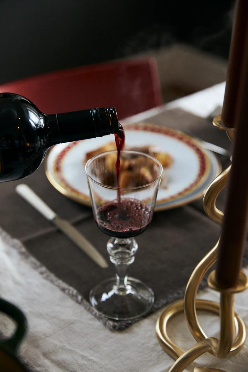 Red wine poured into goblet. Plate with beef casserole, potatoes, fork, and knife. Golden candle holder visible.