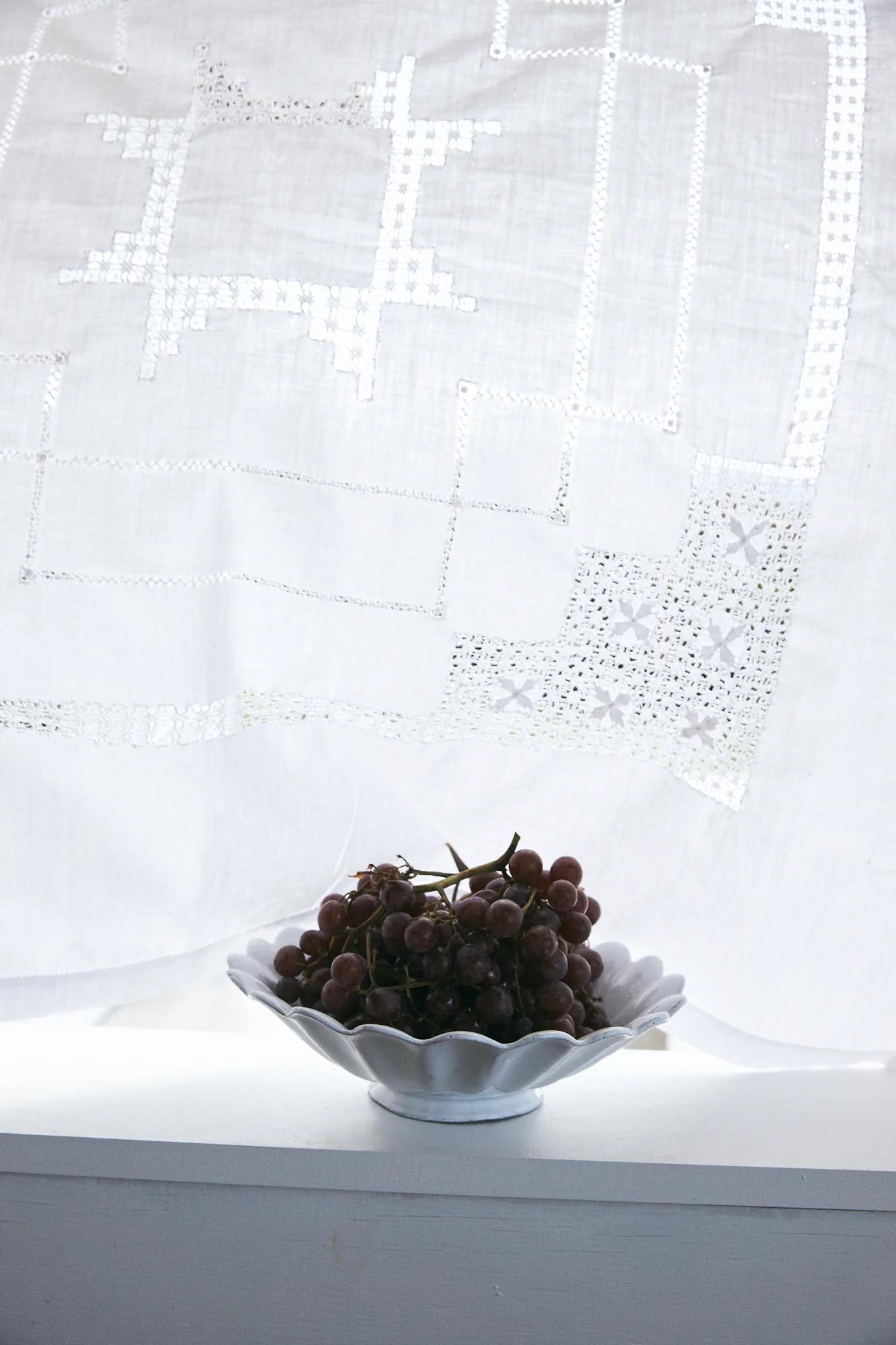 Red grapes in scalloped white bowl on white surface. Lace curtain with geometric patterns hangs behind, diffusing light.