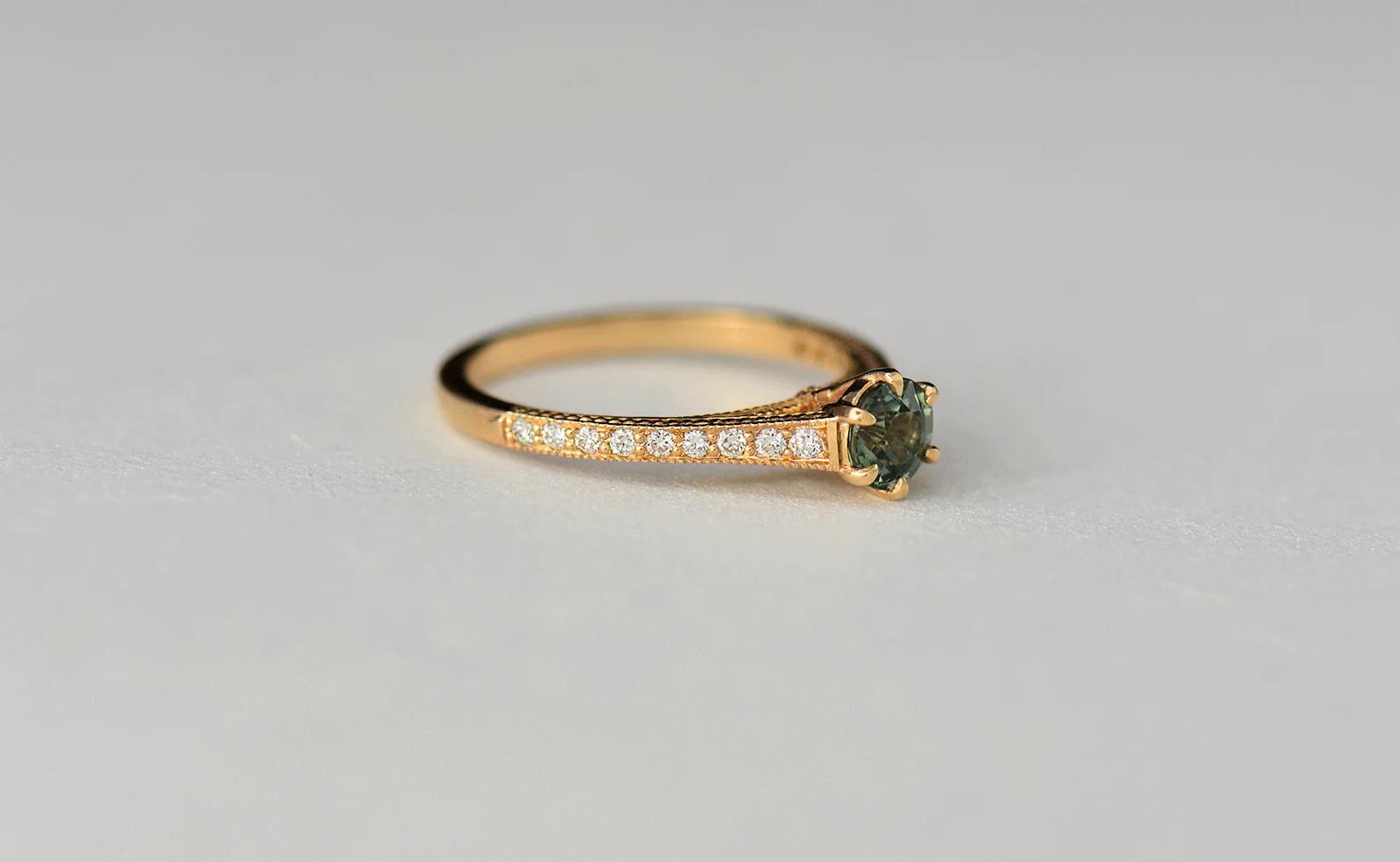 Gold engagement ring with green gemstone and side diamonds. Intricate band detailing. Displayed on light grey background. Charlotte Penman-inspired elegance.