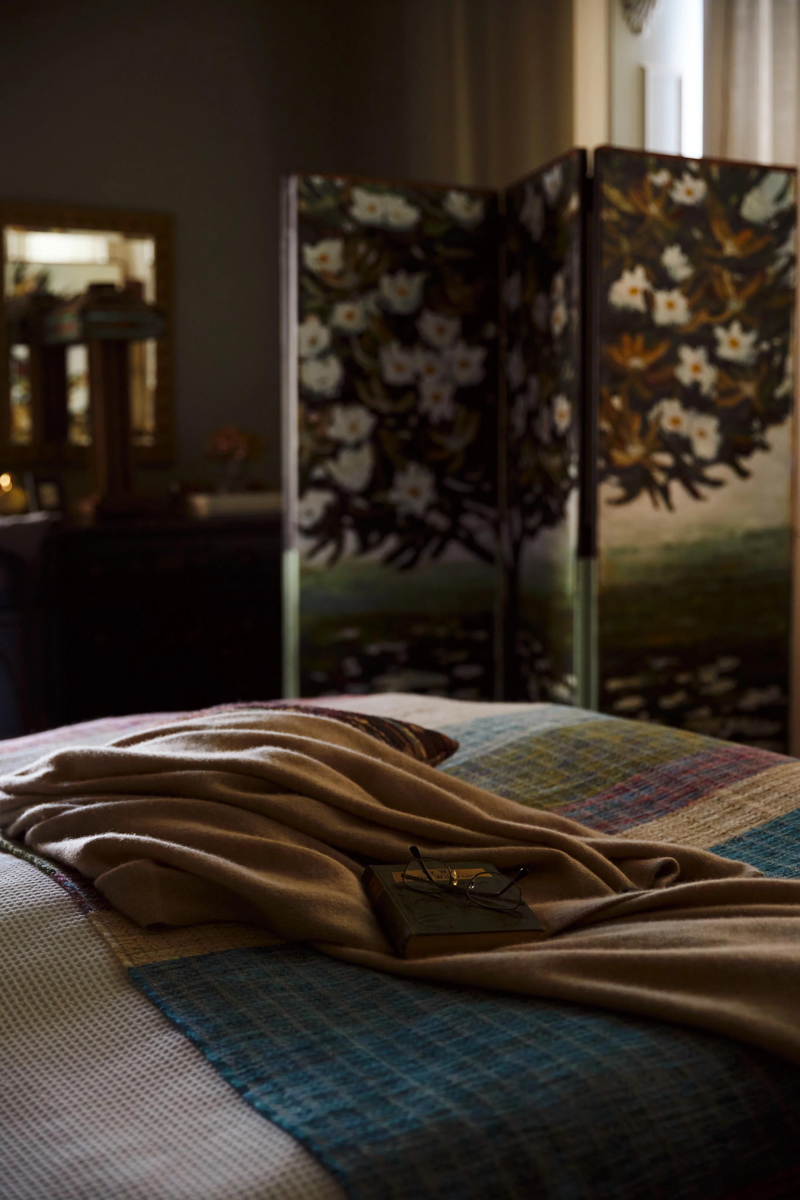 A bedroom with dim lighting features a plaid blanket and beige throw on the bed, a green book and glasses on top, and a wooden screen with floral details in the background.