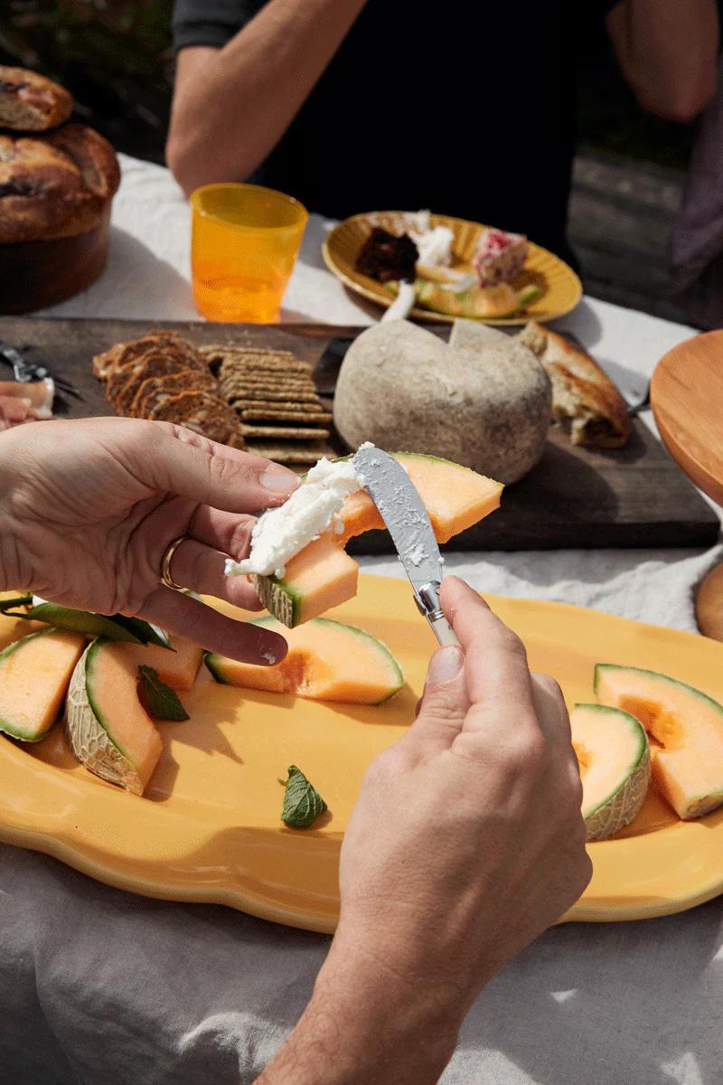 White cheese is spread on a cantaloupe slice on a yellow plate. Crackers, bread, and other dishes are set for a summer outdoor meal.