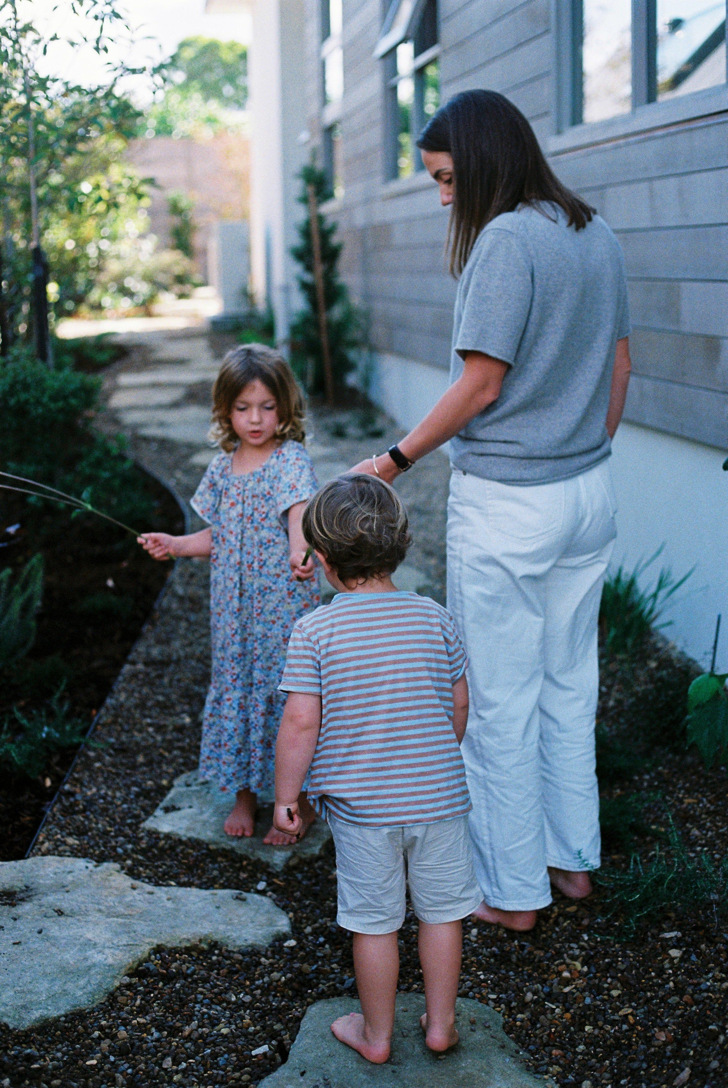 Woman holds hands with young boy in striped shirt. Young girl in floral dress holds twig. Scene on pebbled path near house.