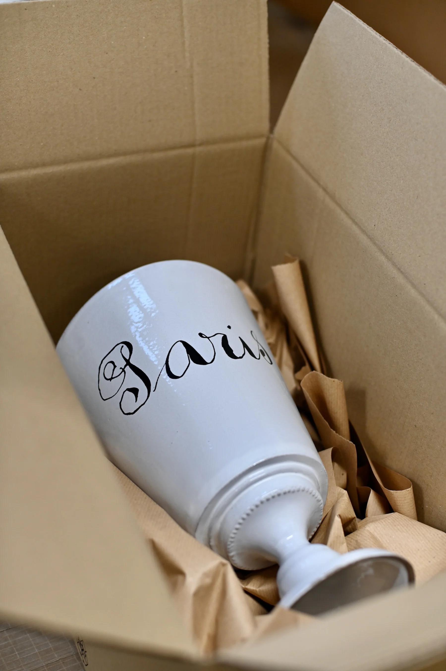 White ceramic vase with "Paris" in black script, partially wrapped in brown paper inside open cardboard box. Black power cord visible.