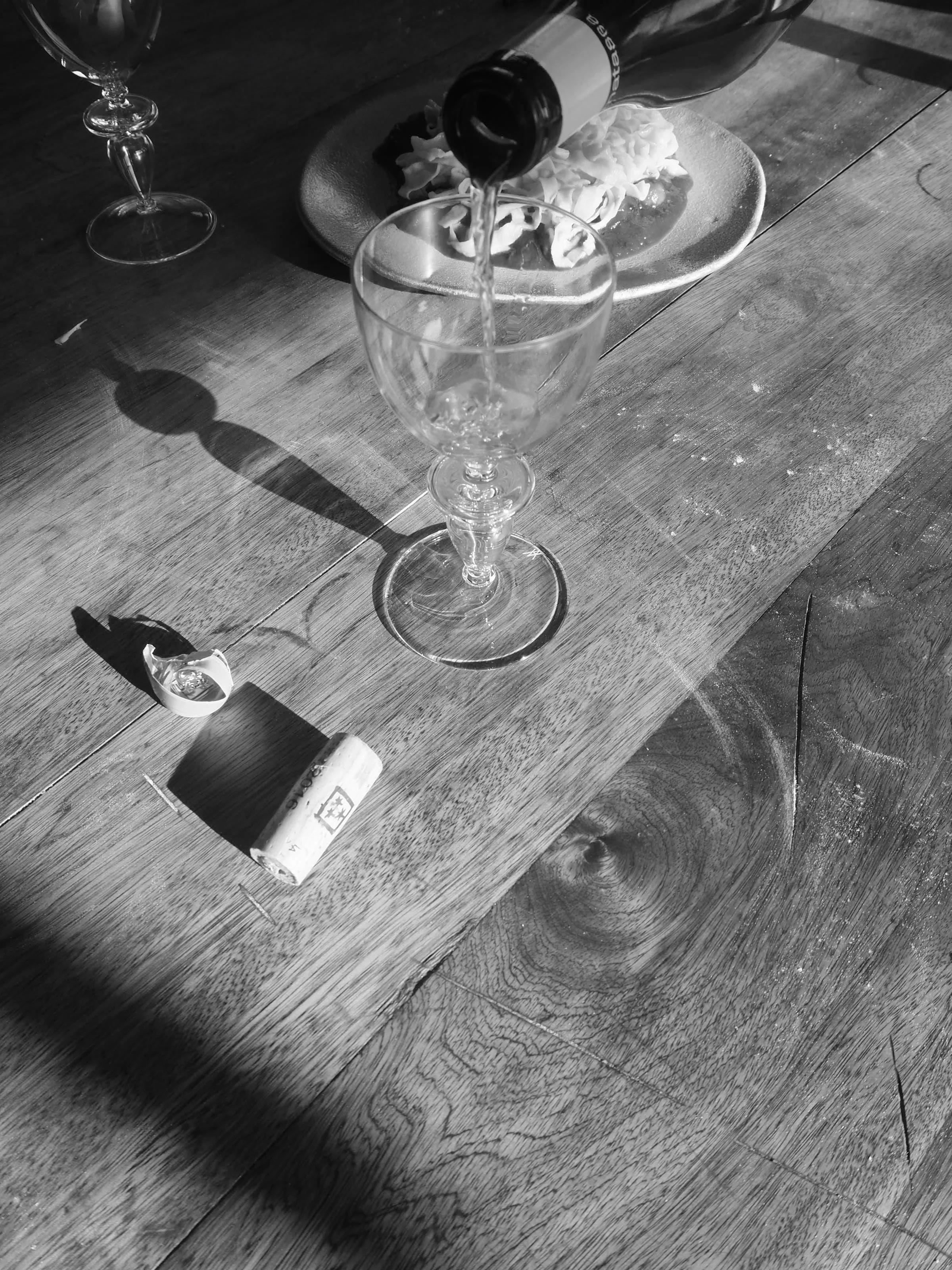 Black and white image: Champagne poured into glass. Cork on wooden table with food plate in background. Sunlight casts shadows across scene.