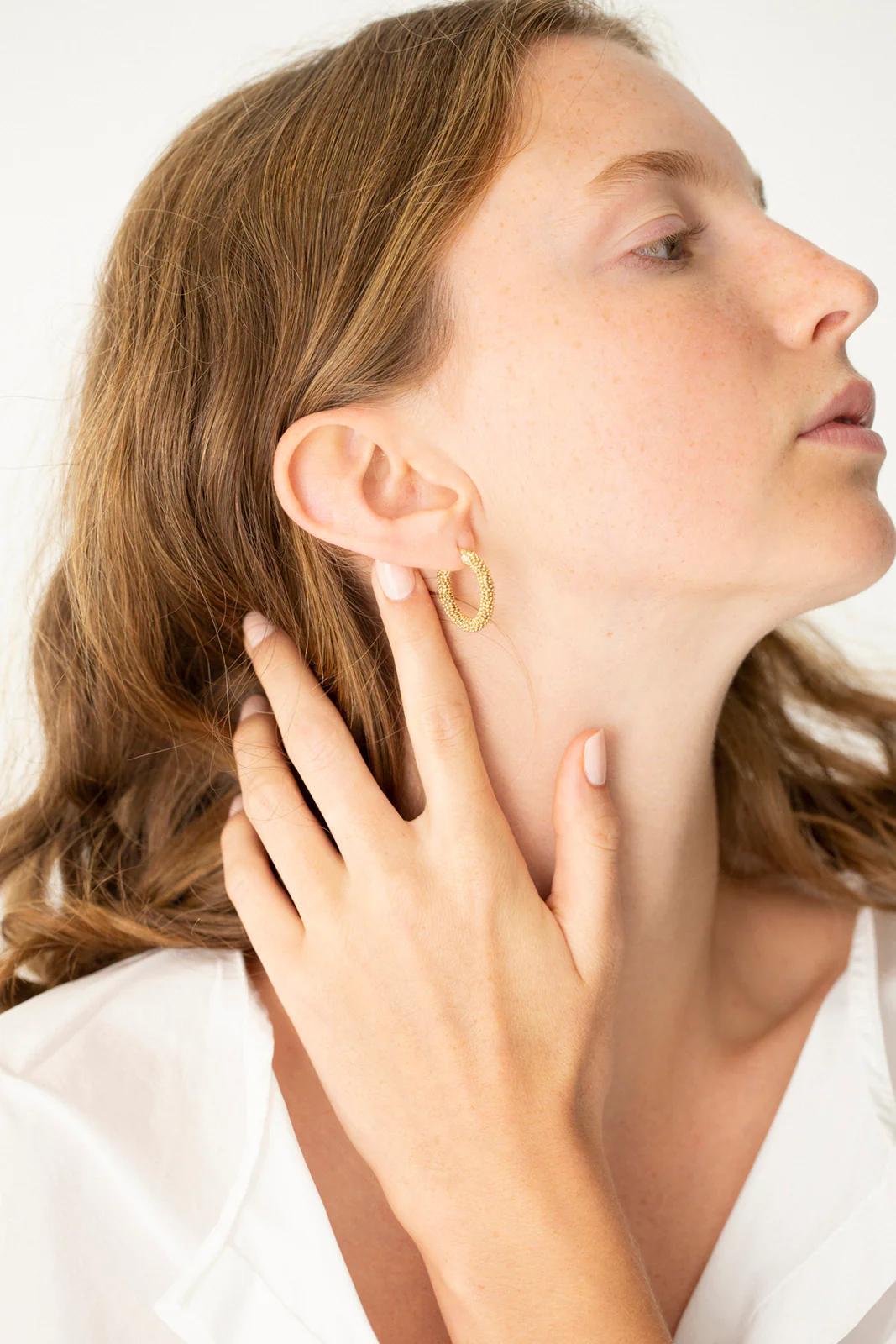 Side profile of person with long wavy hair. Charlotte Penman gold hoop earring. White garment, touching ear gently. Plain white background.