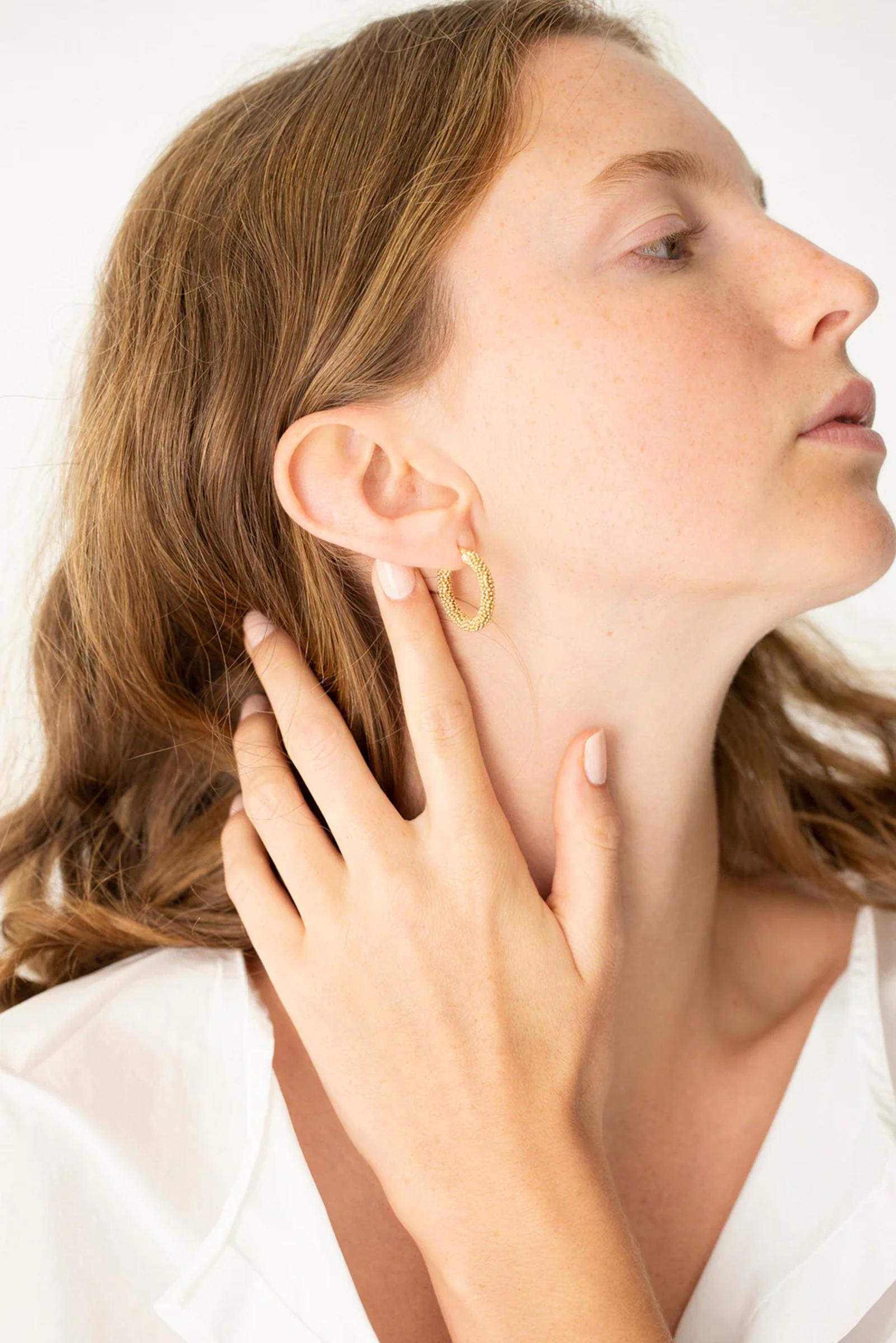 Side profile of person with long wavy hair. Charlotte Penman gold hoop earring. White garment, touching ear gently. Plain white background.