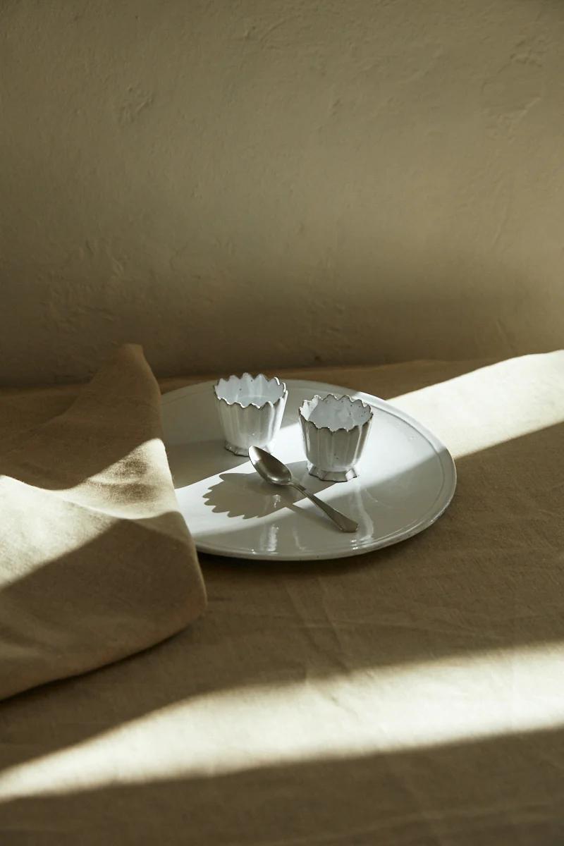 Soft-lit beige table with white plate, metal cups, and spoon. Partially covered by cloth. Warm shadows evoke serene morning ritual.