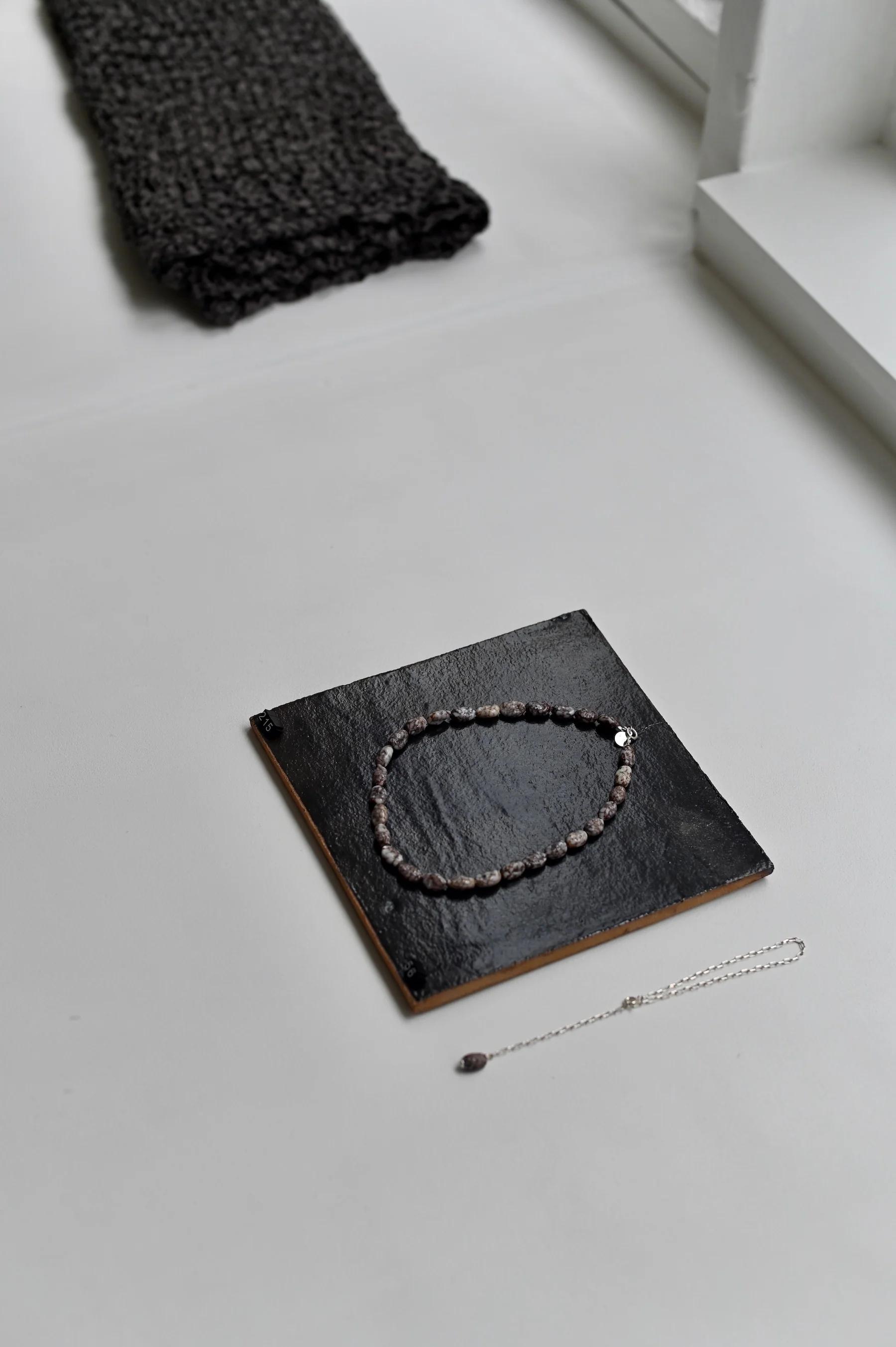 Dark beaded necklace on black platform beside silver bracelet with charm. White surface, dark fabric near bright window.