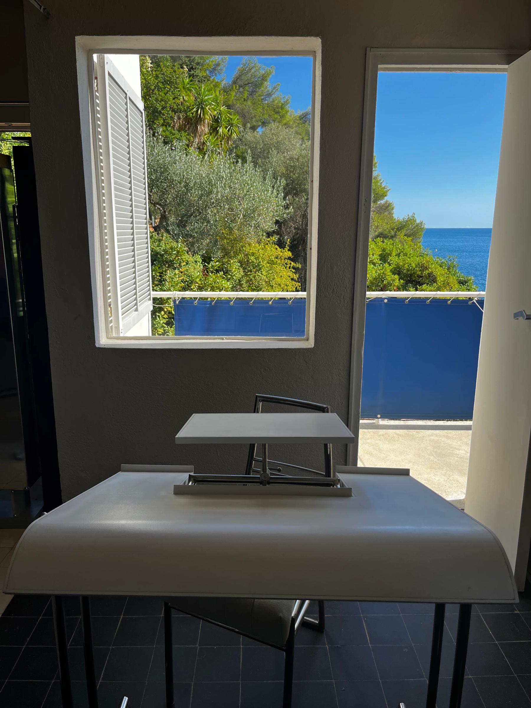 Sunlit room with drafting table and chair. Open window and door reveal green landscape and blue sea.