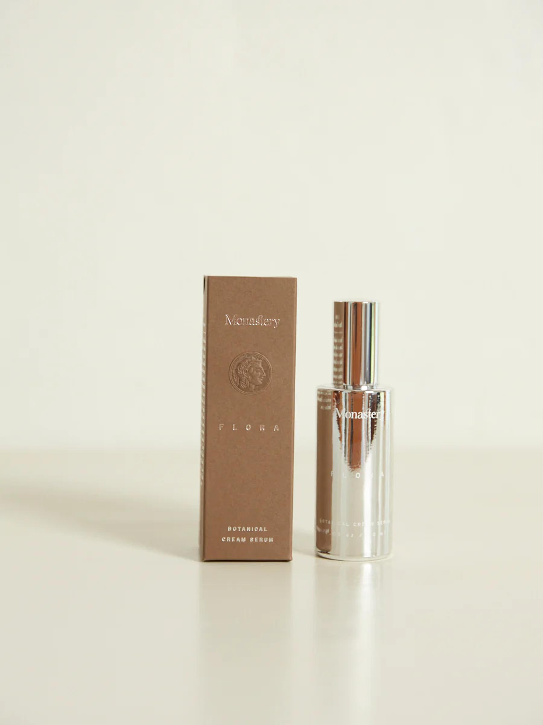 Silver bottle of Monastery Flora Botanical Cream Serum next to its brown box on light surface against neutral background.